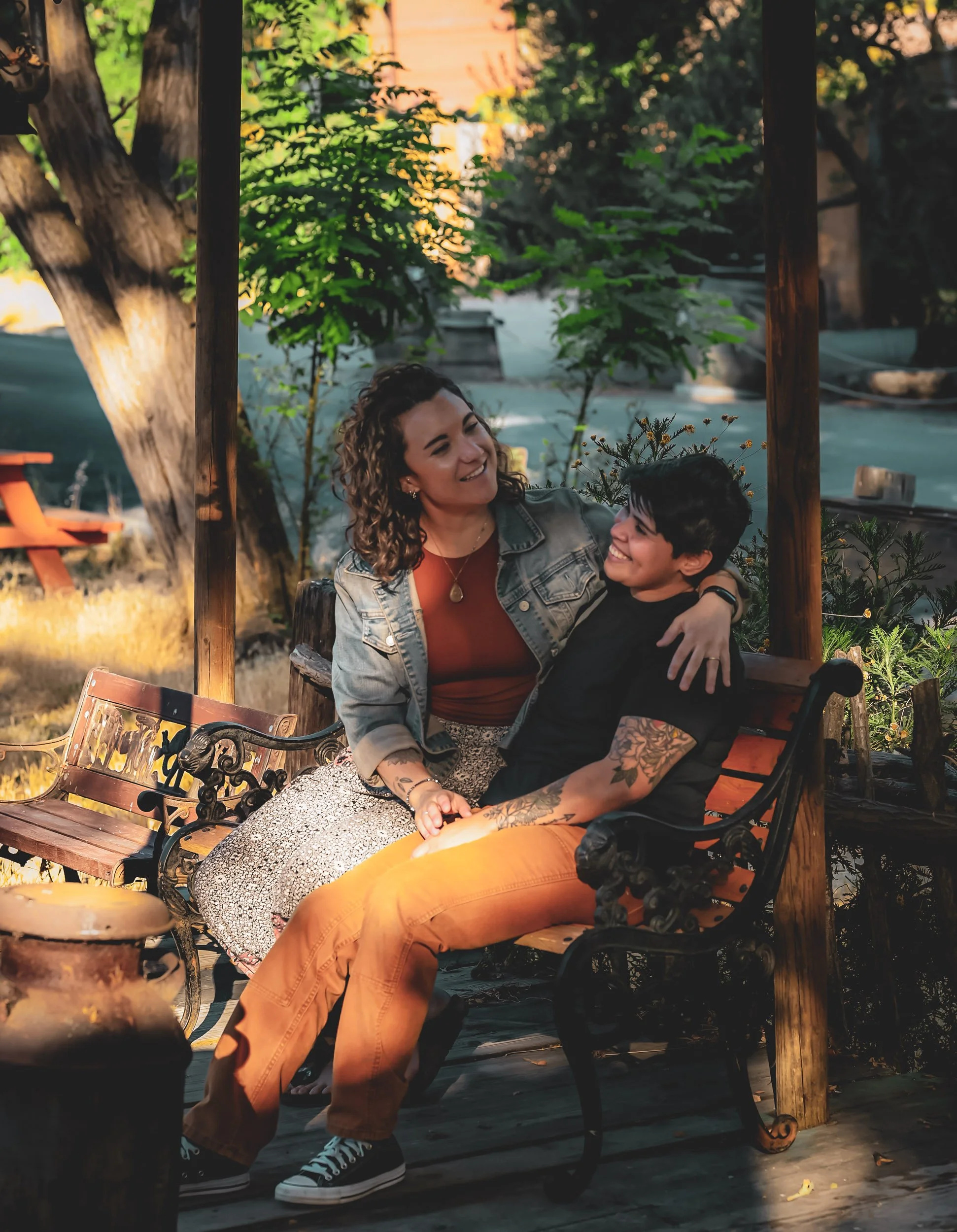 A queer couple sitting on a park bench, smiling and enjoying each other's company, during a fall afternoon in a green outdoor setting.