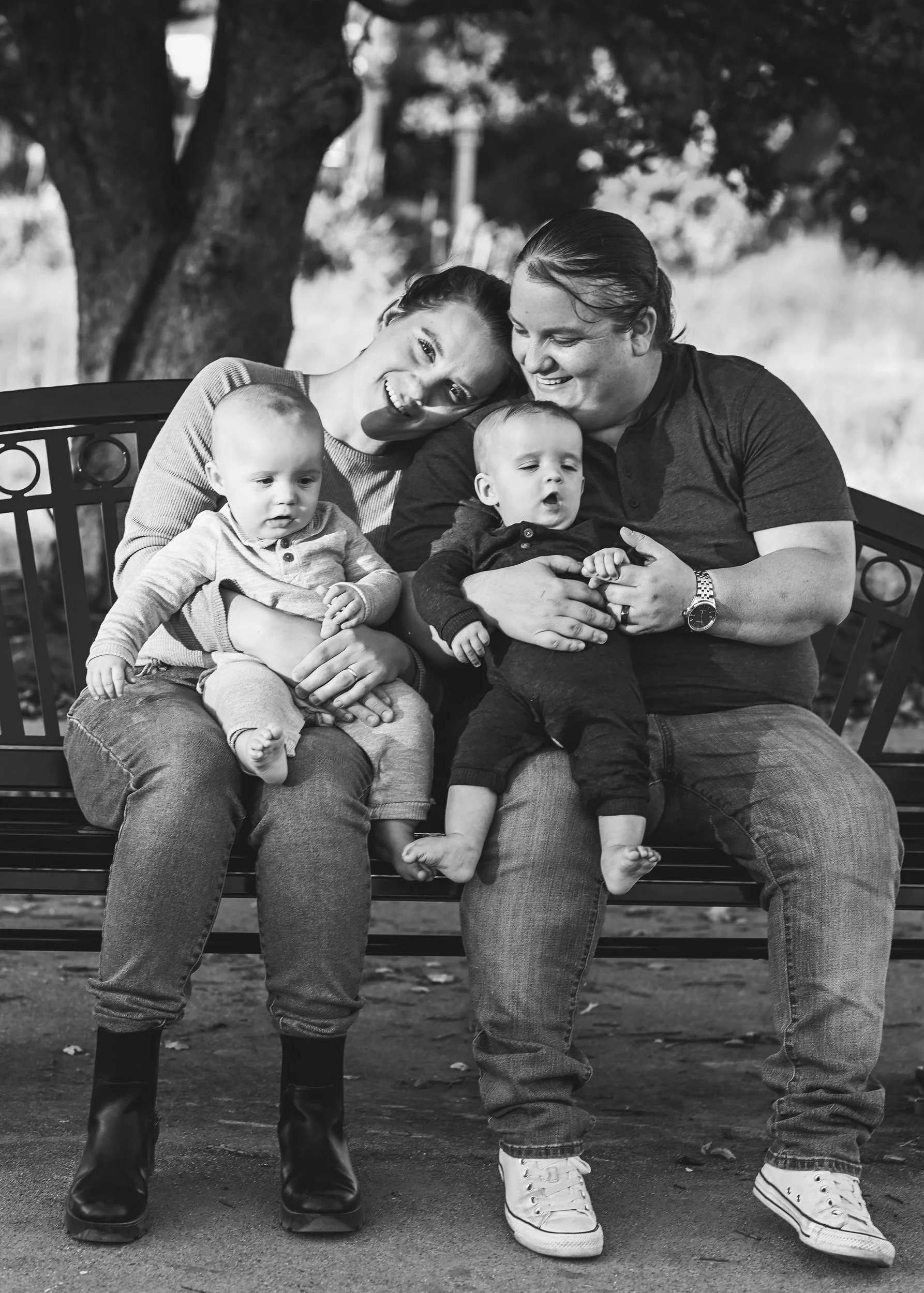 A black-and-white photo of a smiling lesbian couple with two young children sitting on a park bench under a large tree. The children are sitting on the parents' laps, all enjoying a moment of connection together.