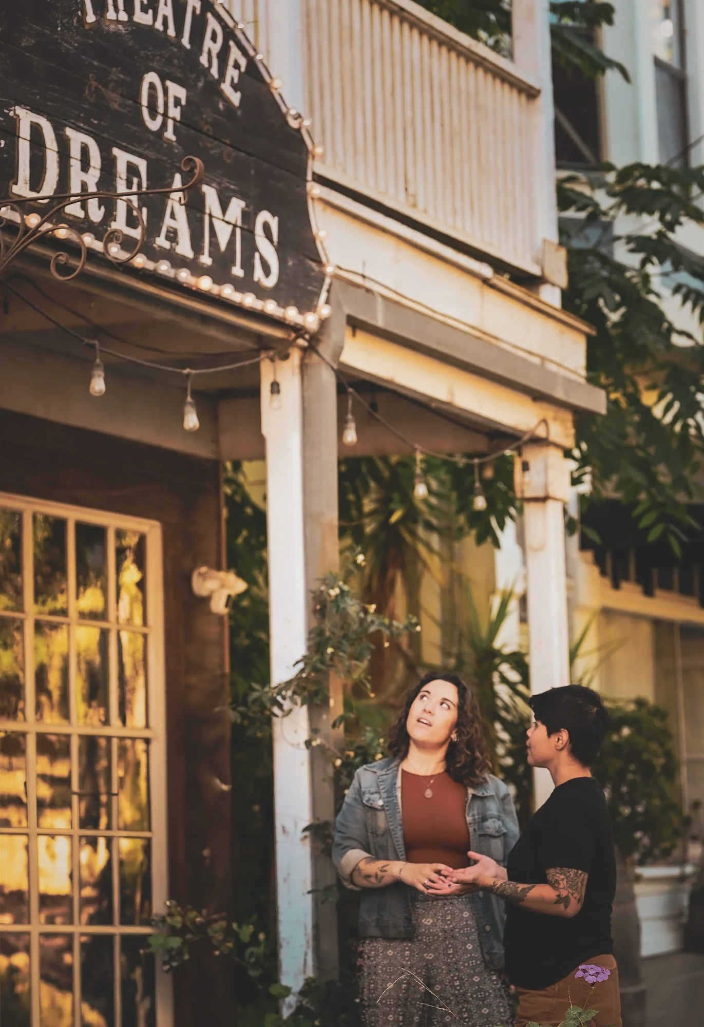 Two women standing outside near a building with a sign that reads 'Theatre of Dreams'.  The couple is talking while a cozy, candid photo is taken.