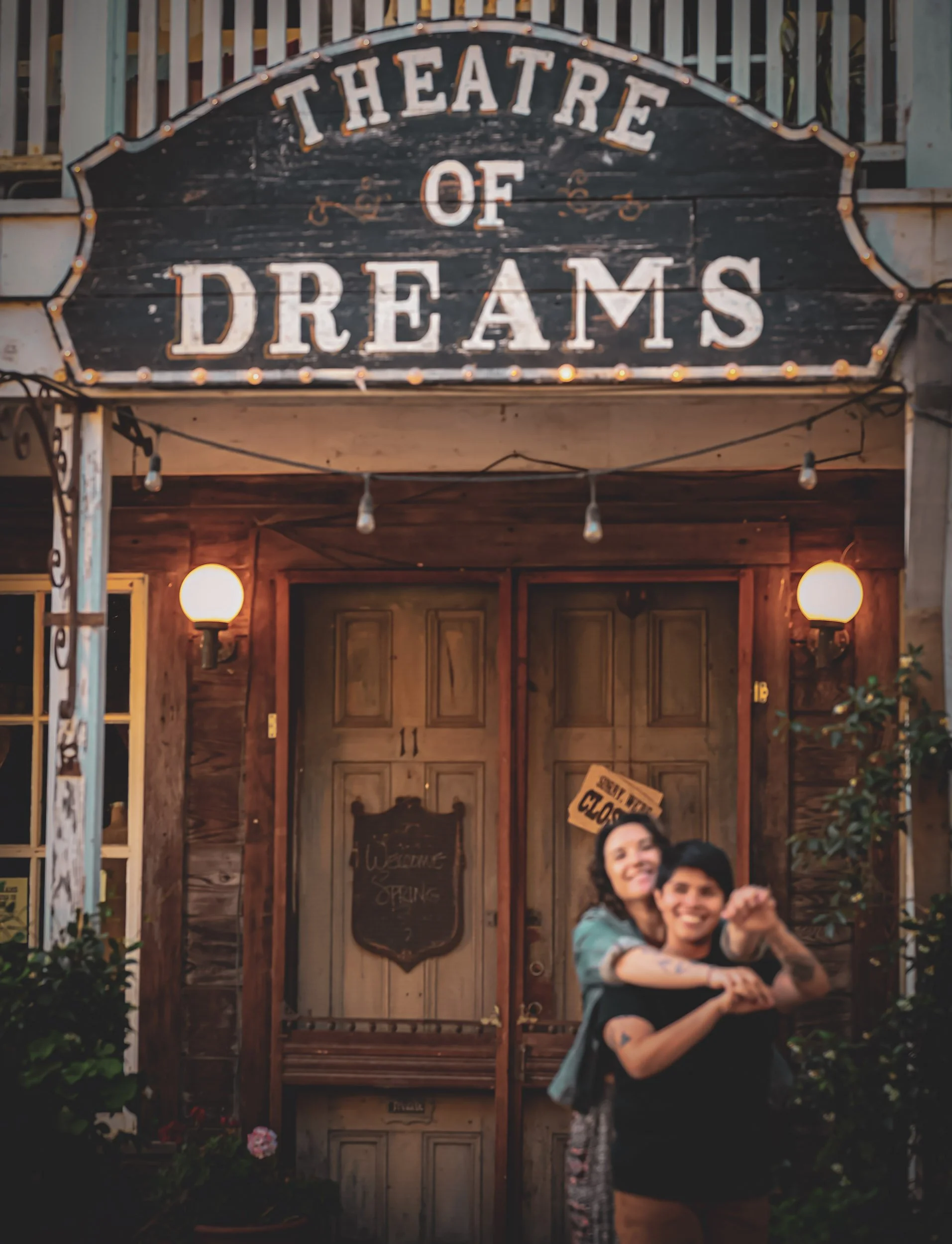 The image shows a wooden theater sign that reads 'THEATRE OF DREAMS' above a door. In front of the door, two smiling people, a lesbian couple, are hugging and looking toward the camera.