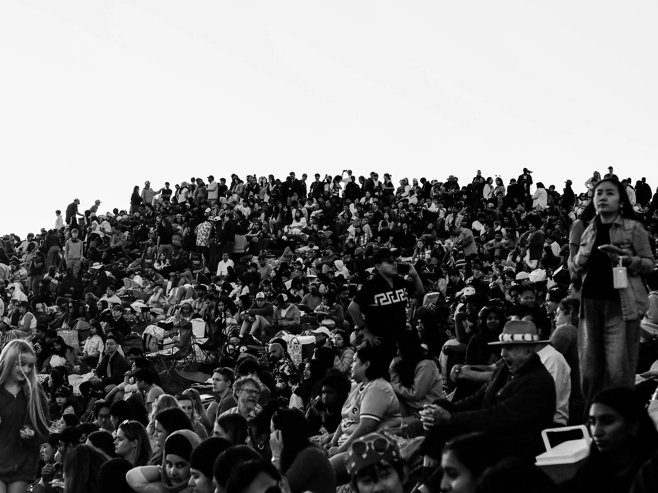 A large crowd of people gathered outdoors, densely packed and sitting or standing on a terraced area, with a clear sky overhead.