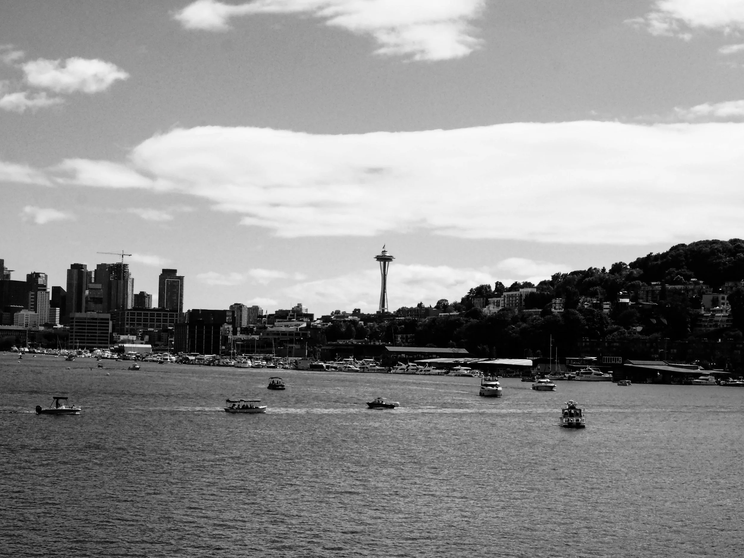 A black and white photo of San Francisco skyline with the Space Needle in the distance and a body of water with boats in the foreground.