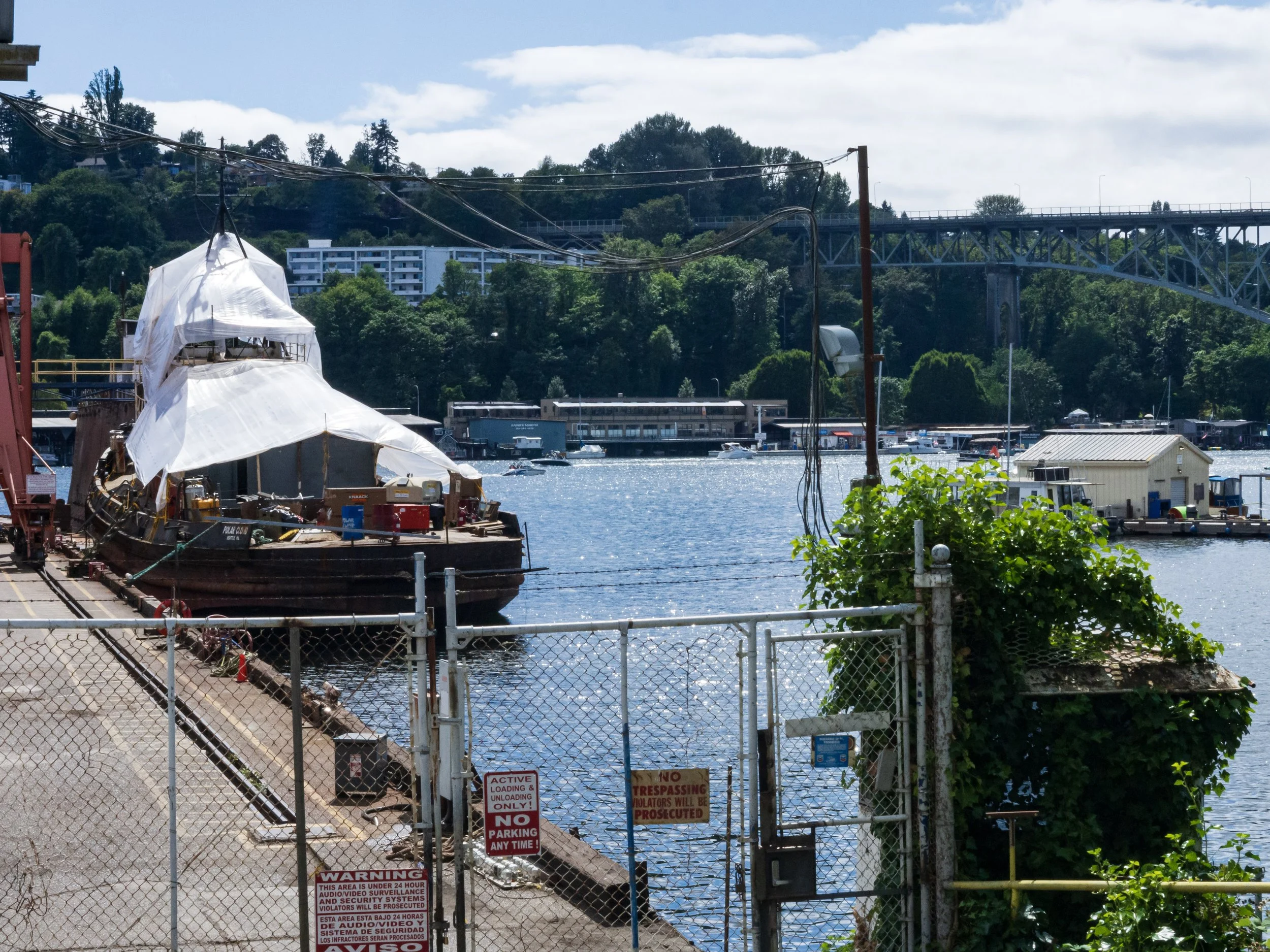 A marina with a boat covered and others docked, a fence with signs, and a bridge over a waterway with buildings and greenery in the background.