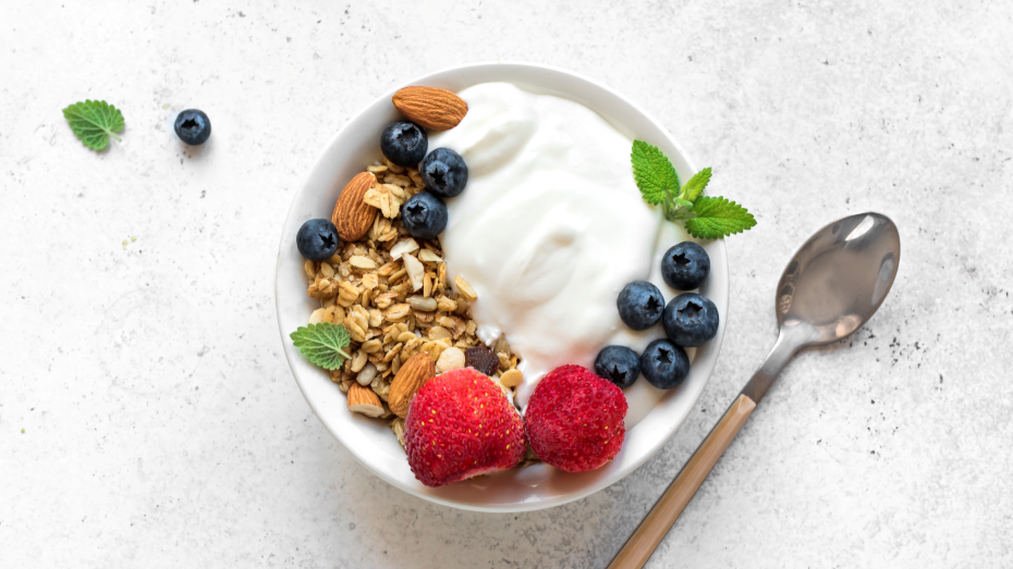 En hvit bolle med yoghurt, granola, blåbær, jordbær og mynteblader. En skje ligger ved siden av.