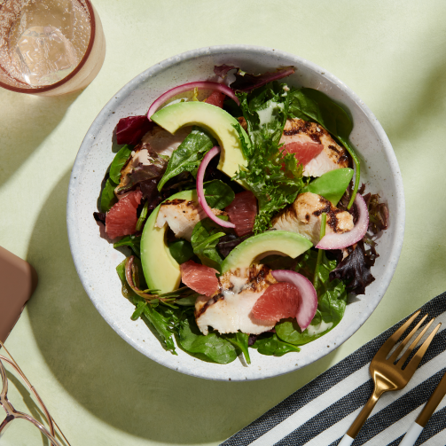 En frisk salat med avocado, rødløk, og tunfisk på en grønn bakgrunn, med et glass vann og bestikk til høyre.