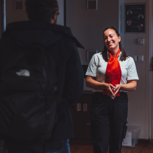 En kvinne i uniform med rødt skjerf snakker med en person med ryggvridt bæreveske i innendørs miljø.