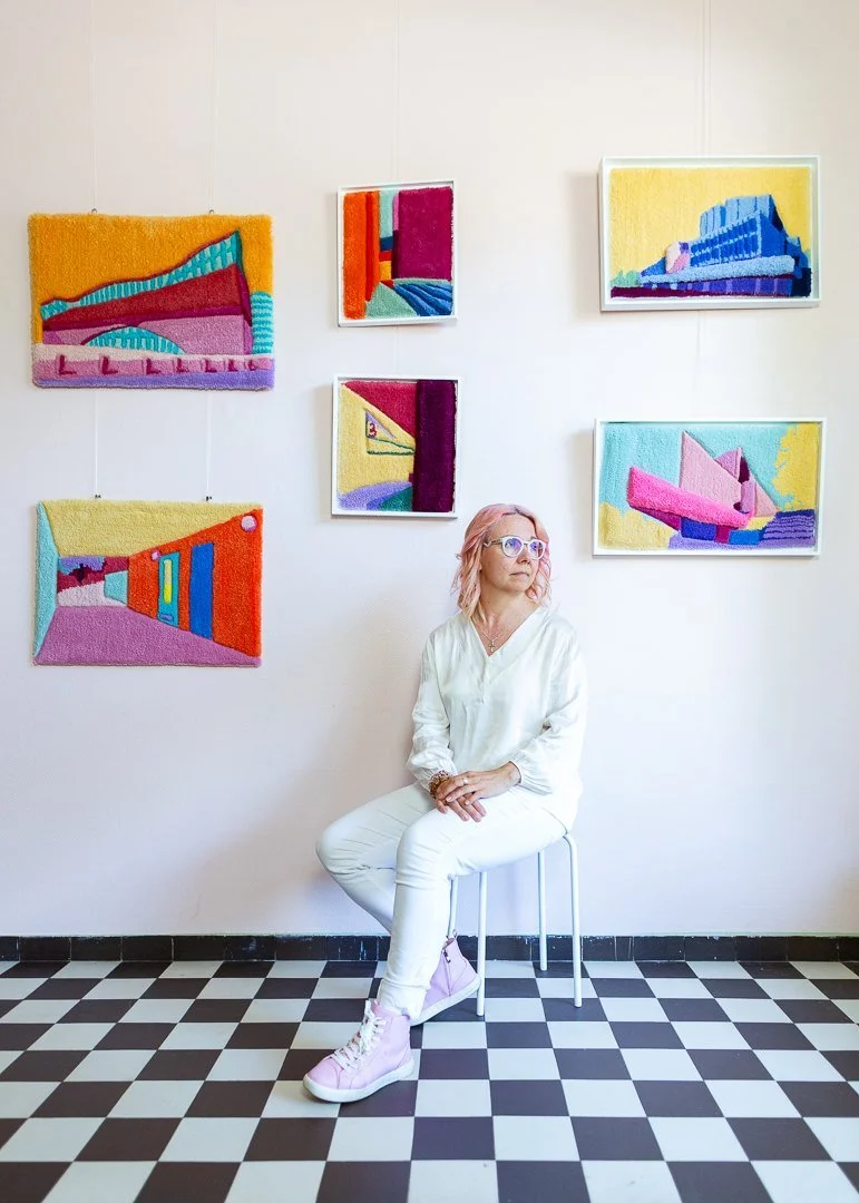 Artist sitting in front of her Helsinki Cityscape Collection, a series of framed tufted textile colourful artworks inspired by Helsinki's architecture and urban landscape.