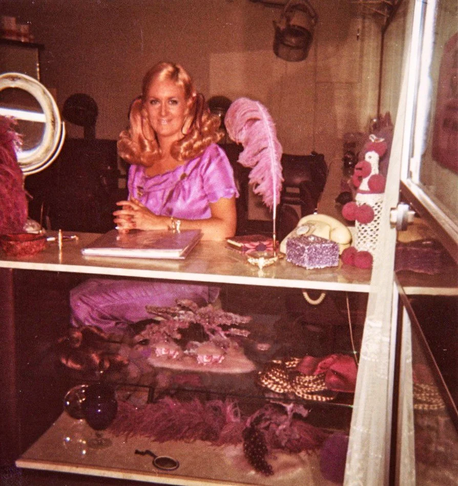 Woman with blonde hair, wearing a pink dress, sitting at a store display counter with various decorative items, including a pink feather quill, a block of soap, and a plush Santa Claus decoration.
