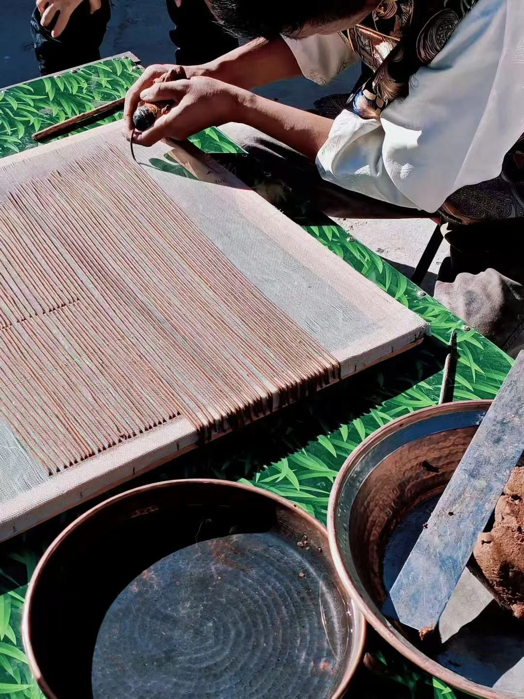 A person working on weaving a pink fabric using a traditional loom, with copper bowls and a metal sheet nearby on a green leaf-patterned table.