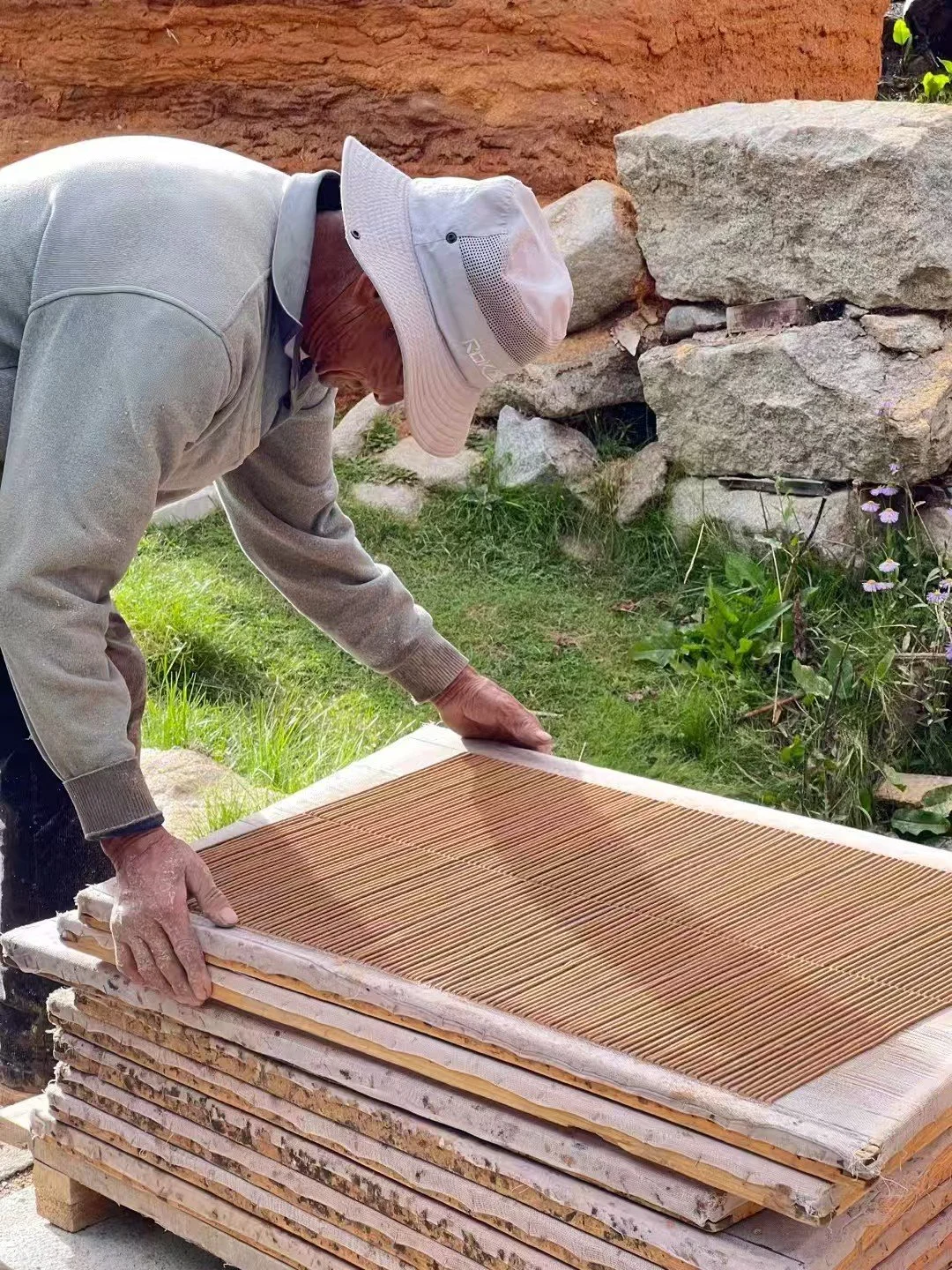 An elderly man wearing a light-colored hat and glasses bends over a stack of large ceramic tiles or stone slabs, outdoors near rocks and greenery.
