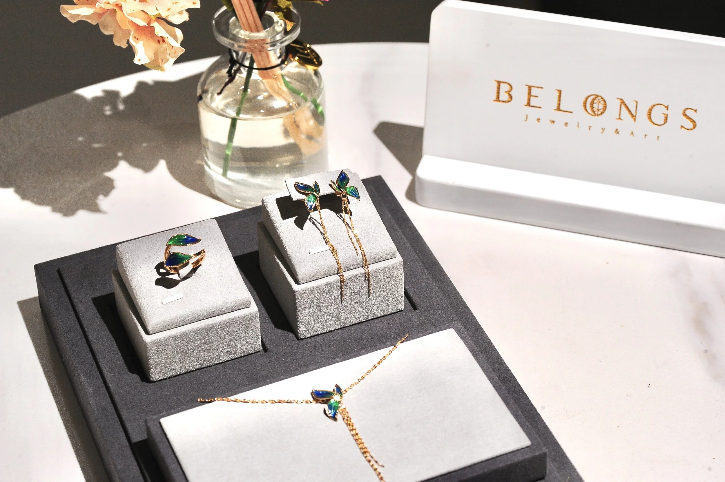 Display of jewelry including a ring, necklace, and earrings, with a vase of pink flowers and a sales sign for Belongs Jewelry & Art in the background.