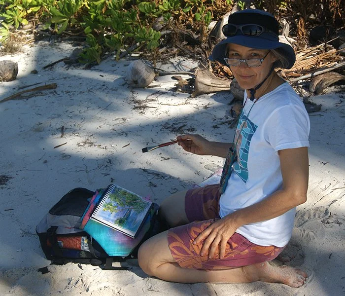 Judith Künzle mit Sonnenhut und Brille, sitzen im Sand am Strand, Pinsel halten, hat Malblock vor sich, umgeben von Pflanzen und Strandgut