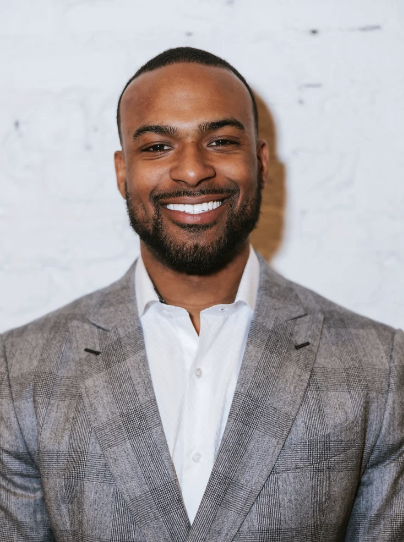 Professional man with a beard, smiling, wearing a gray suit and white shirt, standing against a light-colored brick wall.