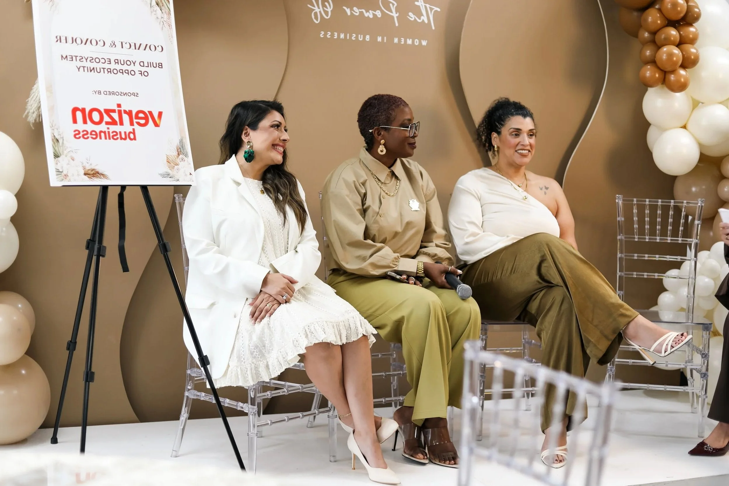 A panel of three women sitting on transparent chairs at an event, smiling and talking. A balloon decoration in neutral colors is visible in the background. A sign on a stand with red and black text reads about a venture business event.