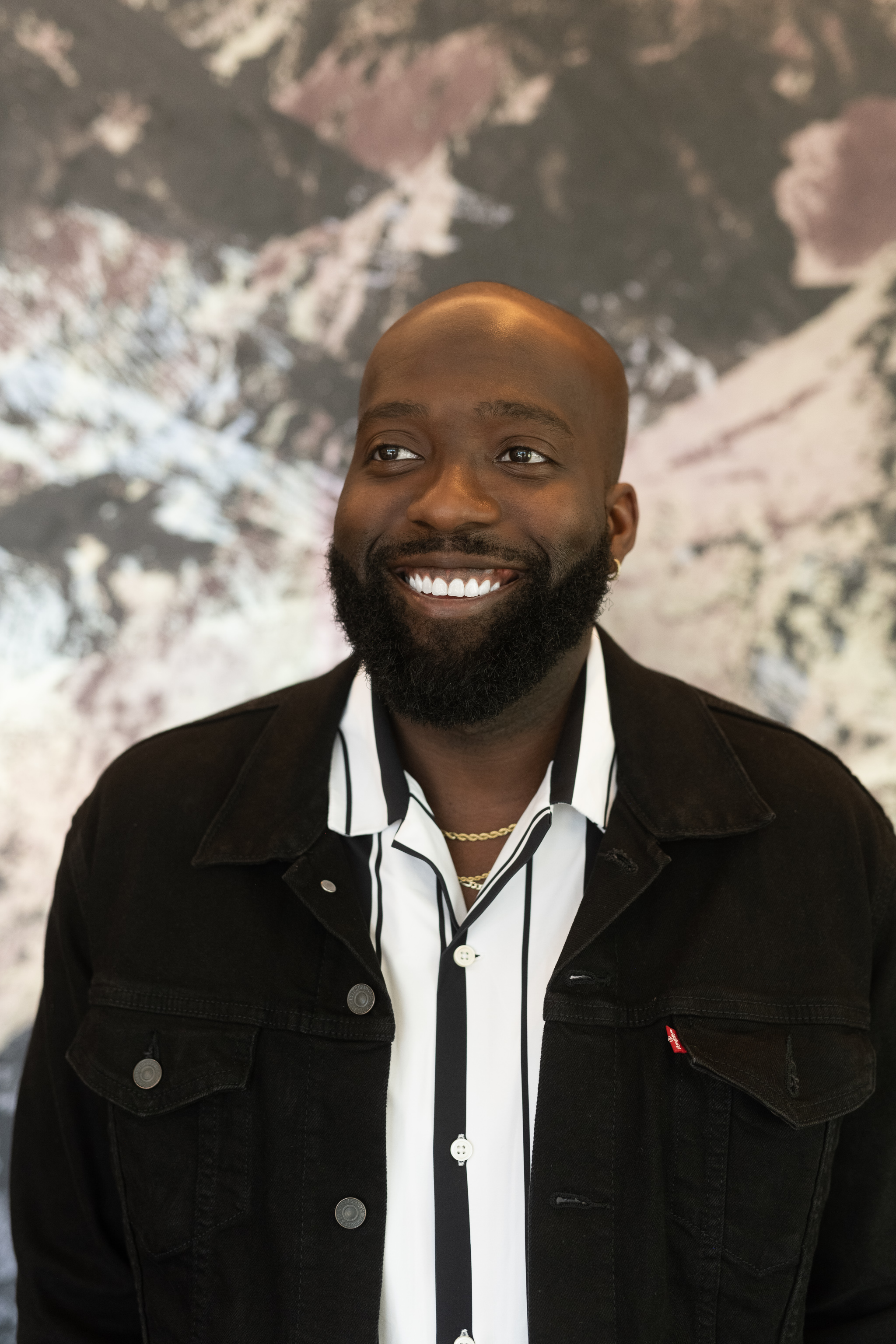 A smiling Black man with a beard wearing a black denim jacket over a white shirt with black stripes, and layered gold necklaces, standing in front of an abstract textured background.