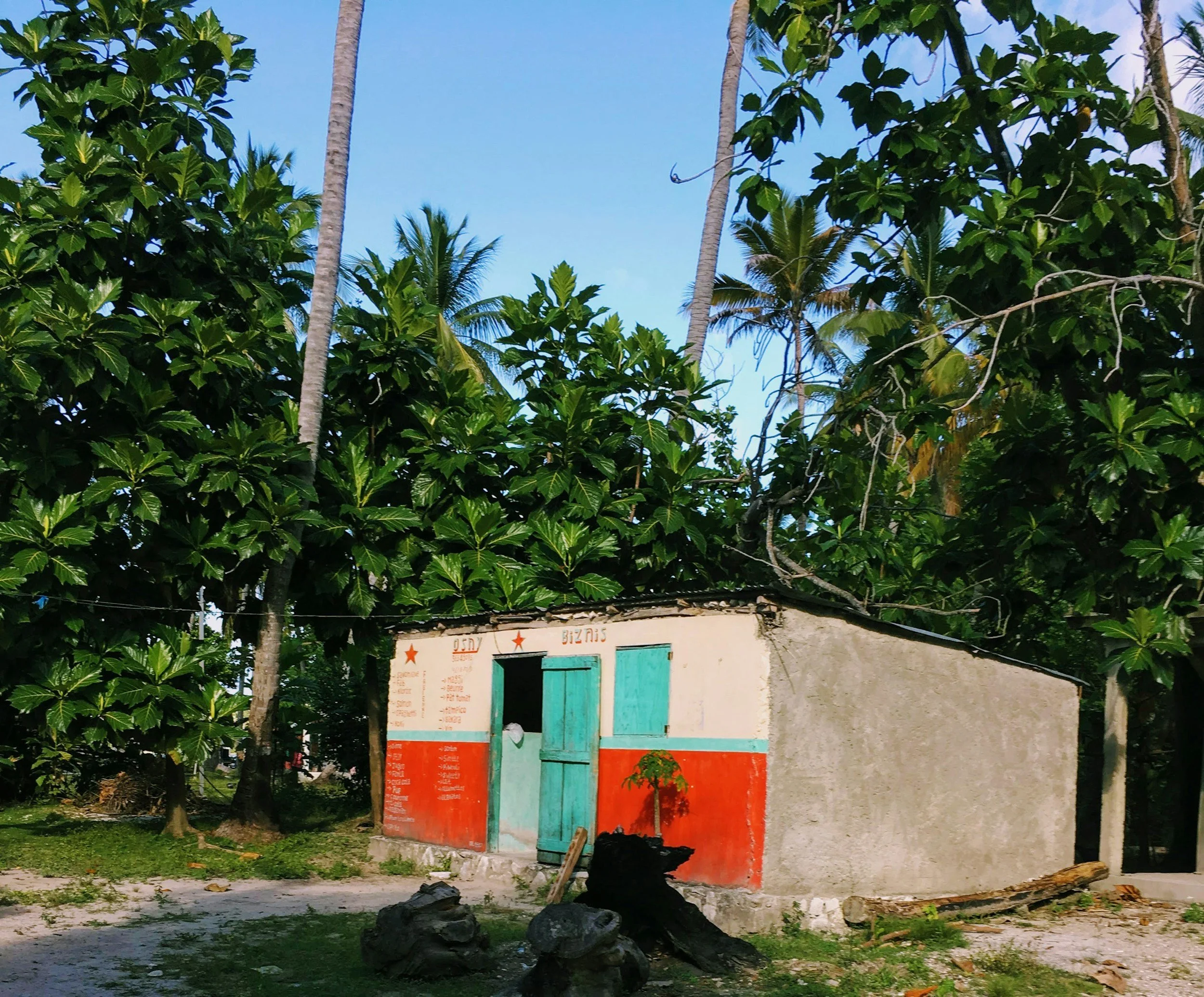 Small white building with turquoise double doors and red signage, surrounded by lush green trees and blue sky.