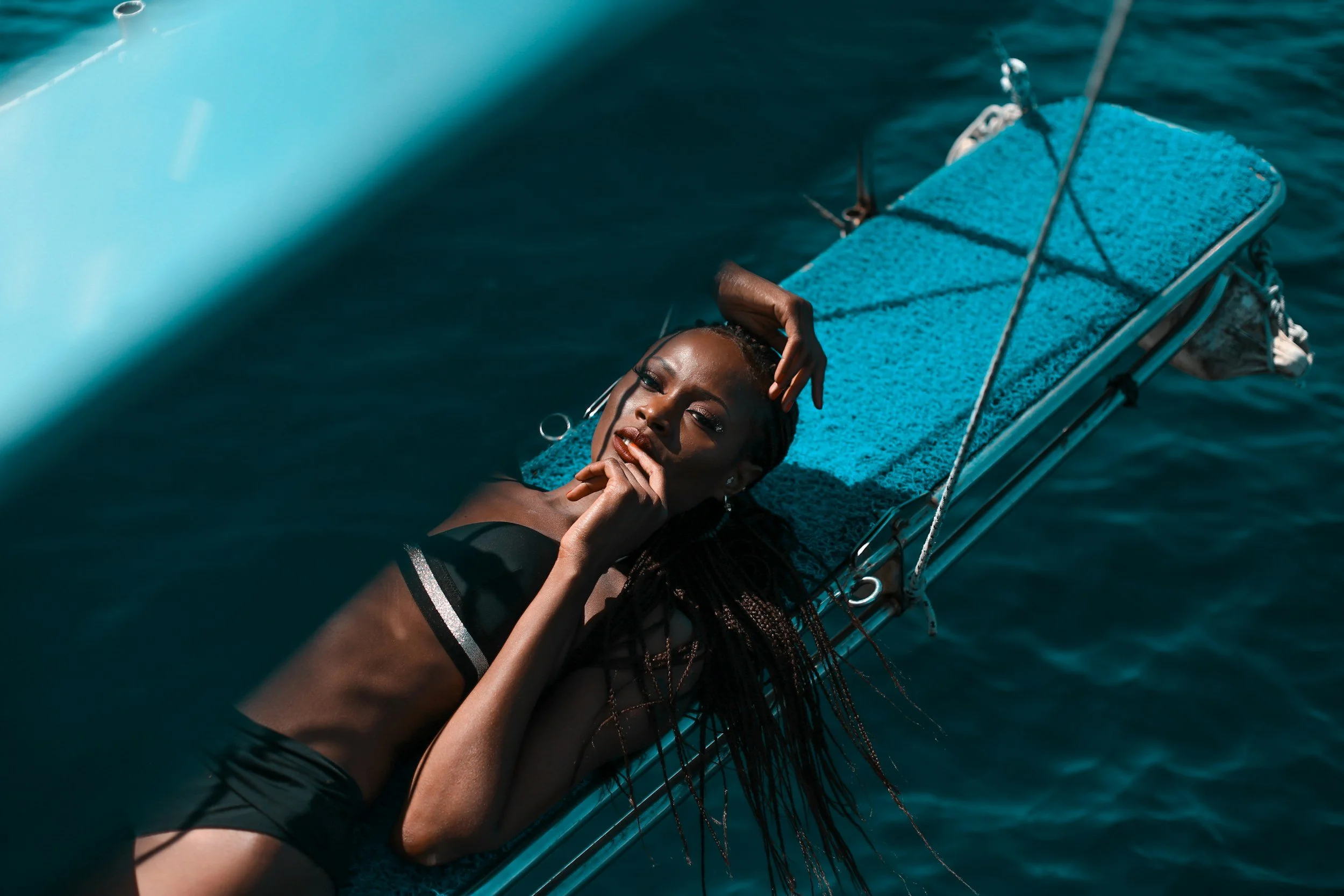 A woman with dark skin and long braids lying on a blue textured bench on a boat, with dark water surrounding her.