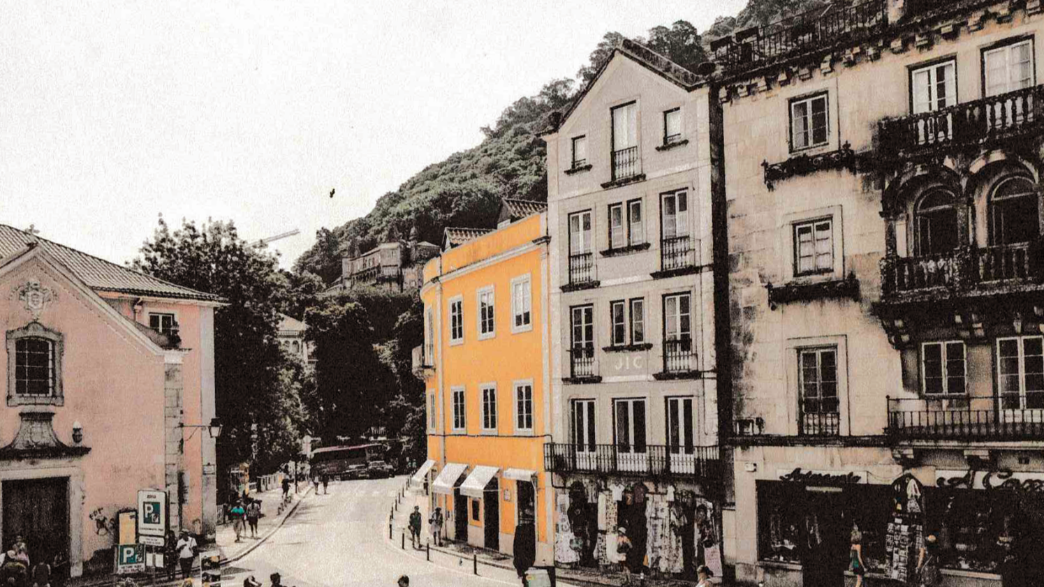 Colorized photograph of a street scene with multi-story buildings, including an orange building with white striped awnings on the ground floor, people walking, and a hillside with trees in the background.