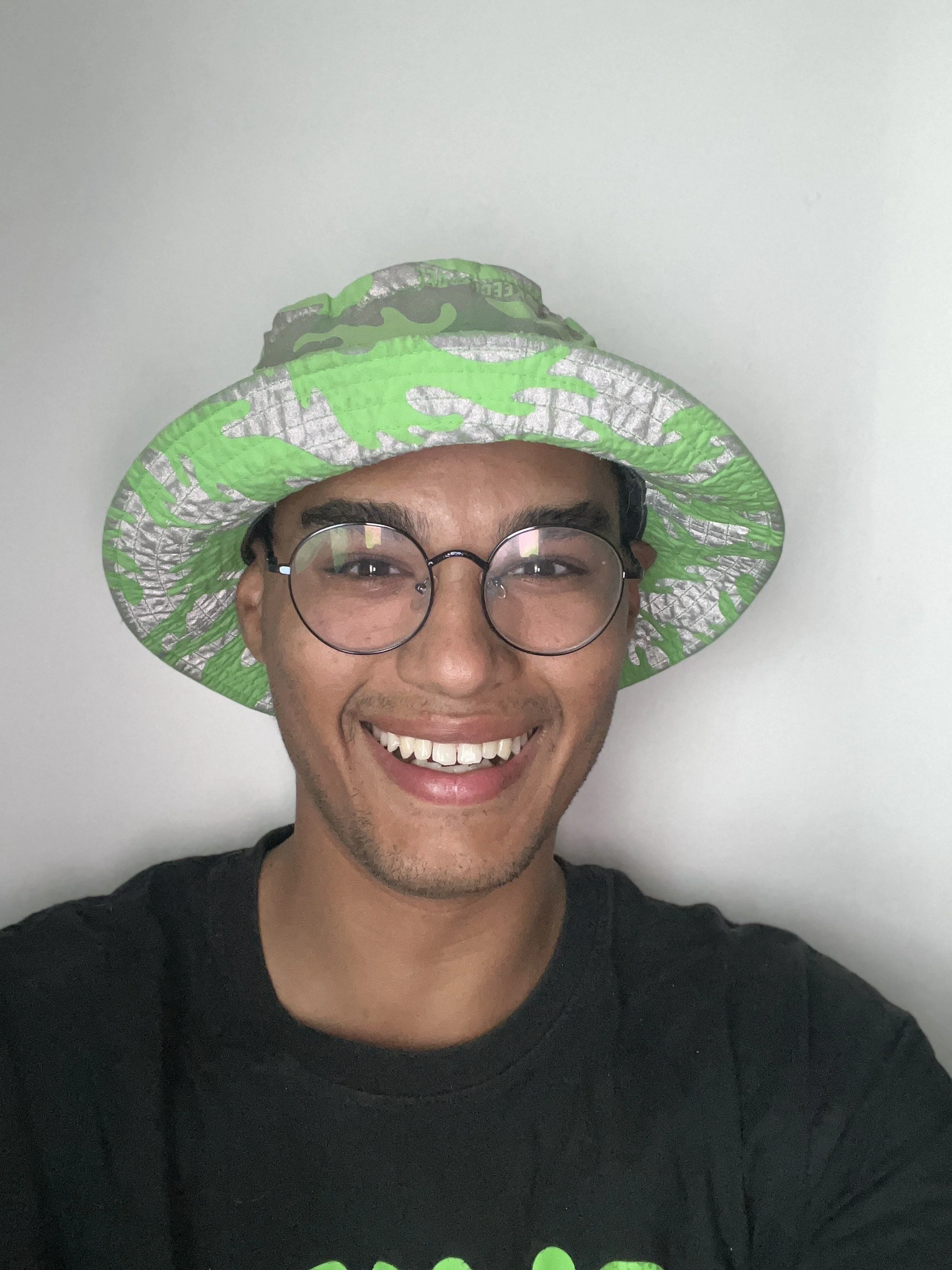 A person with glasses smiling, wearing a green and silver hat with black designs, and a black shirt with green lettering, standing against a plain white wall.