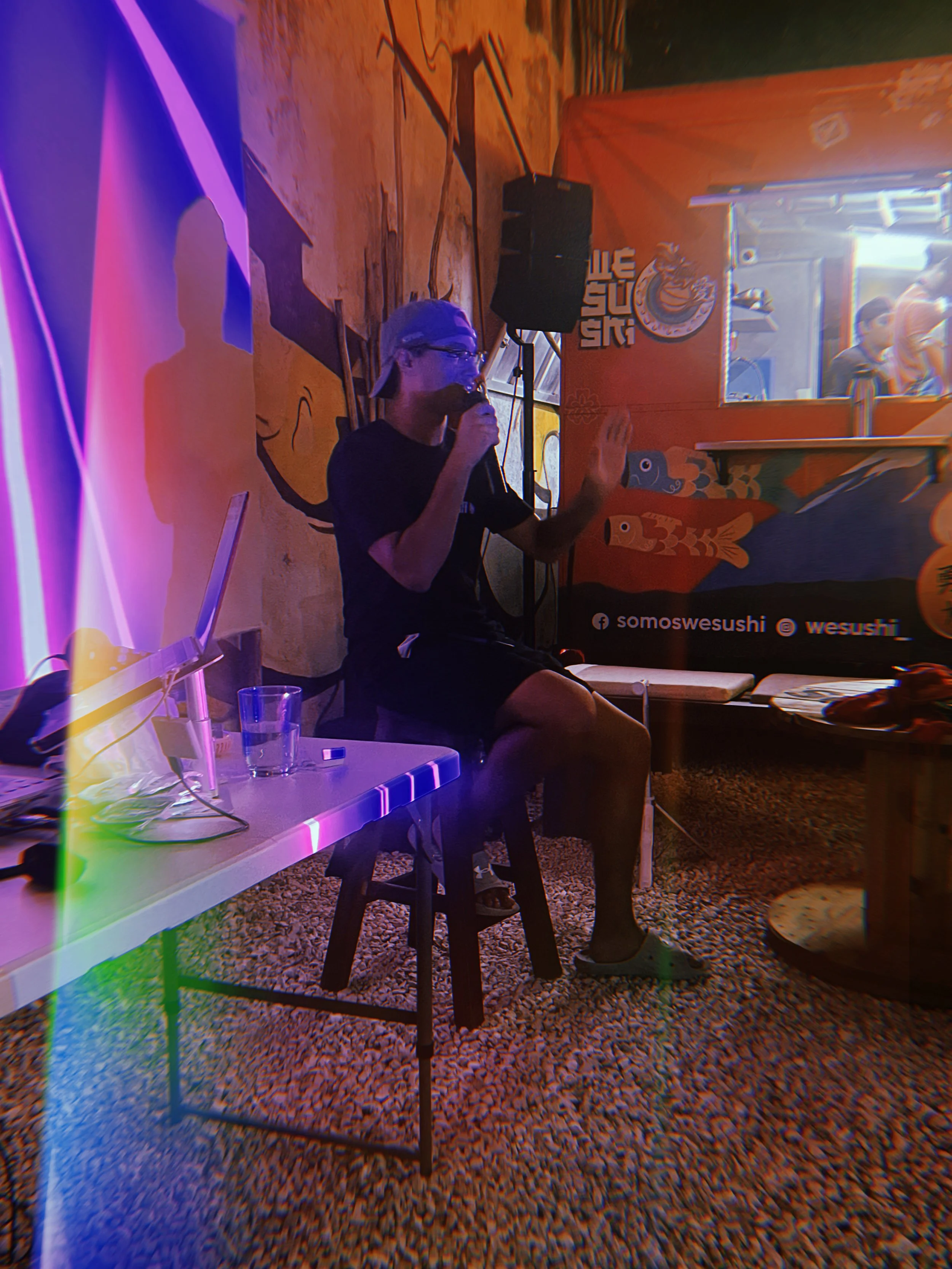 A man with gray hair, glasses, and a black shirt, sitting on a black stool, speaking into a microphone in an indoor setting with colorful decorations, a neon light, and a small crowd in the background.
