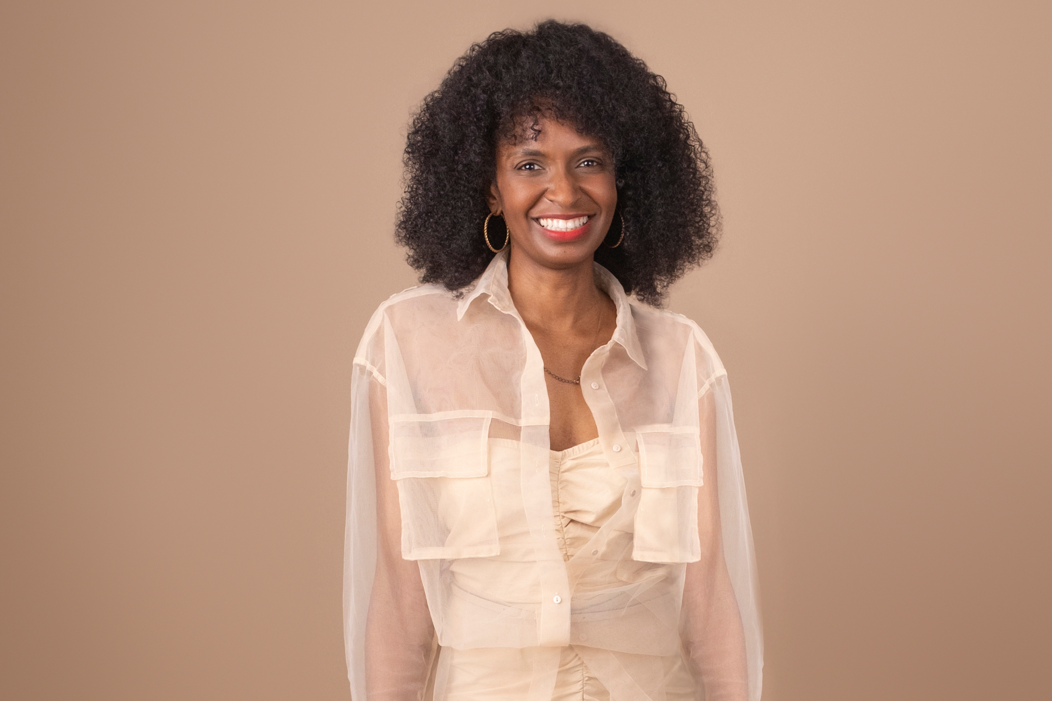 A woman with curly hair wearing a light-colored shirt and hoop earrings, smiling, standing against a plain beige background.