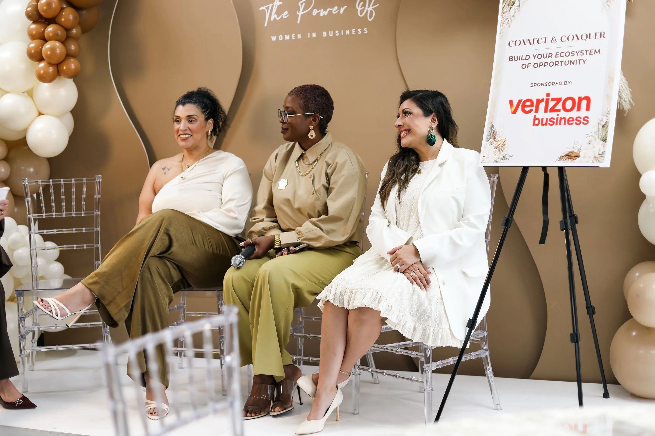 Three women sitting on a stage at a business event, with a sign behind them that reads 'The Power of Women in Business' and 'Connect & Conquer: Build Your Ecosystem of Opportunity,' sponsored by Verizon Business. The stage is decorated with balloons.