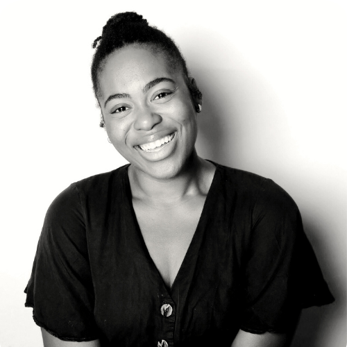 A smiling woman with natural hair styled in a bun, wearing a dark top, against a light background.