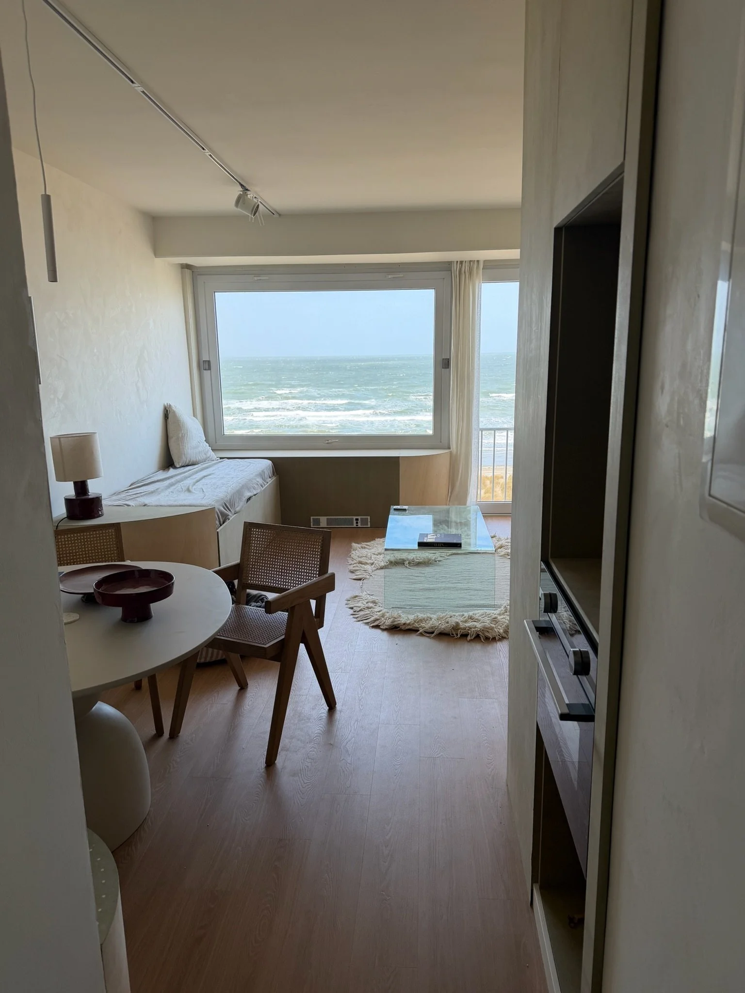 A studio with a large window overlooking the ocean, a small seating area with a sofa and pillows, a coffee table, a wooden dining table with chairs, a lamp, and a bookshelf or cabinet.