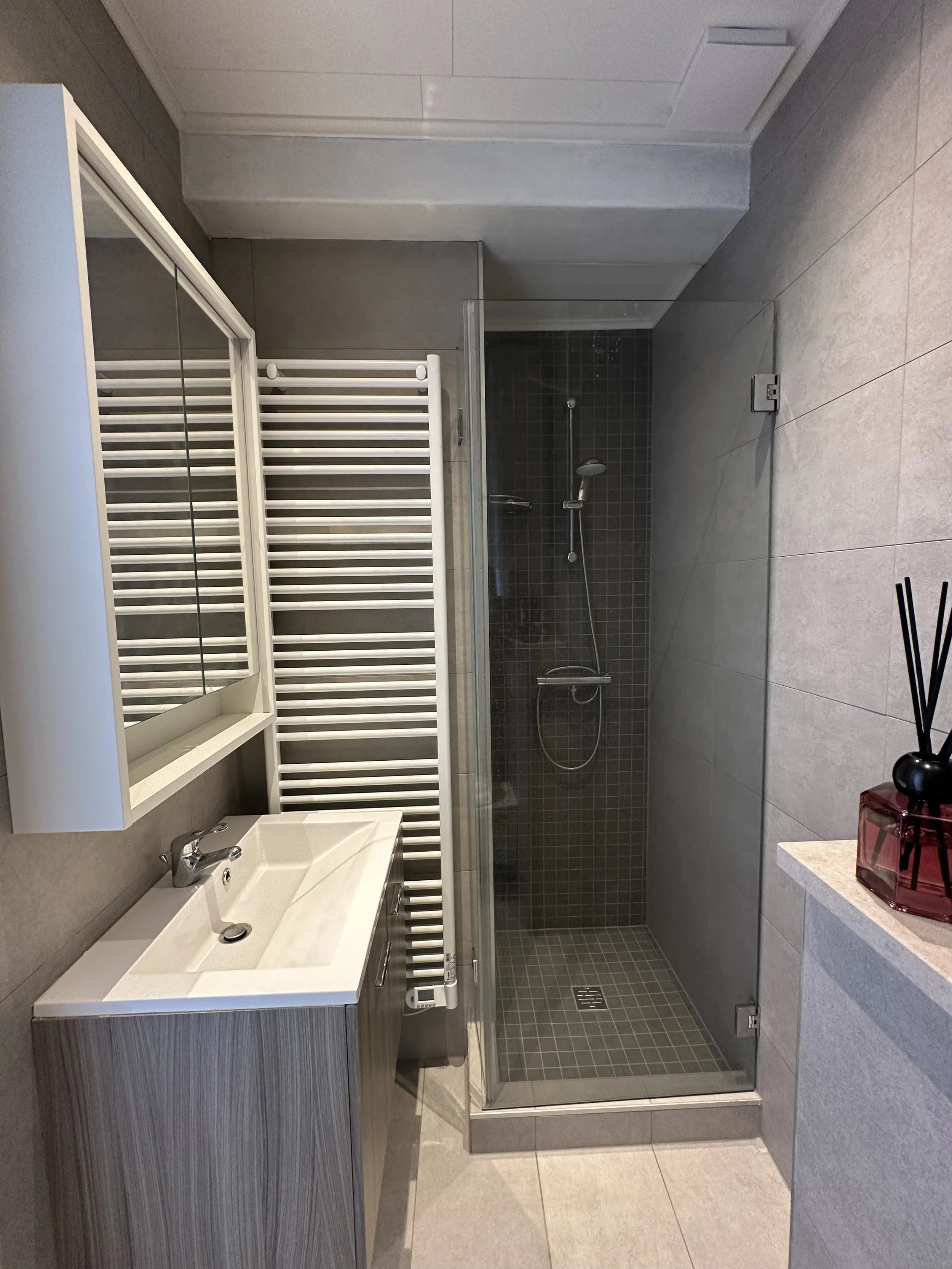 A modern bathroom with a shower area, a white sink with a mirror, and a white towel radiator on the wall. The shower has dark tiles and a glass enclosure. The floor has large light-colored tiles.