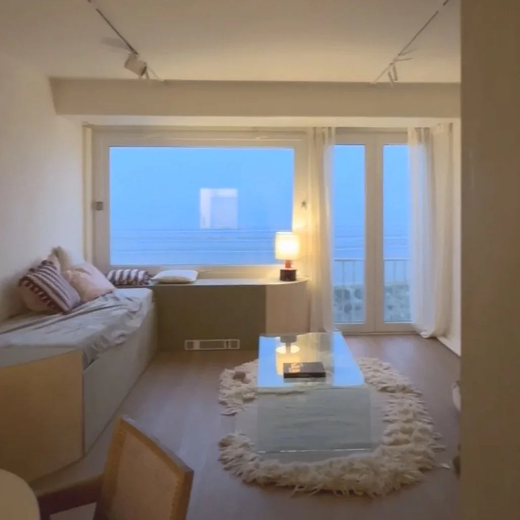 Cozy living room with large window view, white curtains, sofa with striped pillows, glass coffee table on a furry rug, and a small table with a lamp in the corner.