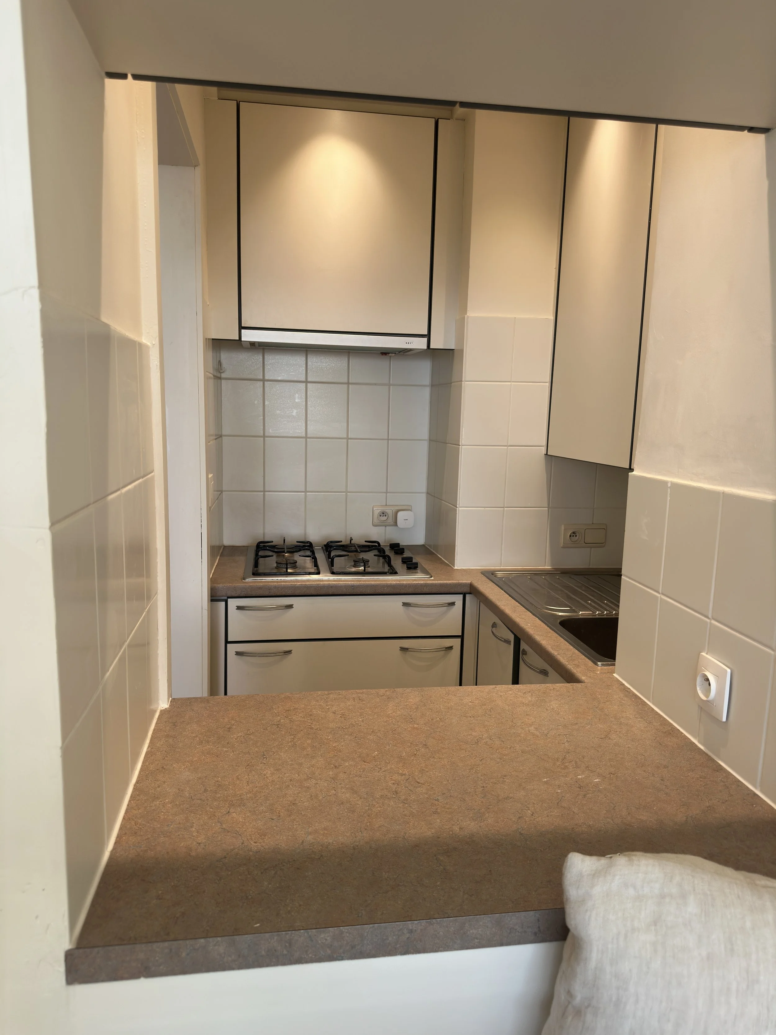 View of a small kitchen with a countertop, a stove, a sink, and upper cabinets. A cushion in the foreground suggests a seating area nearby.