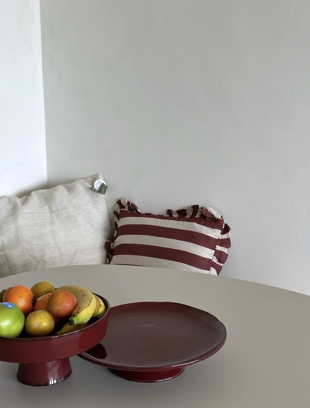 A white round table with two red cake stands, one holding various fruits including apples, bananas, and oranges, and the other empty. In the background, two pillows are on a sofa against a plain white wall.