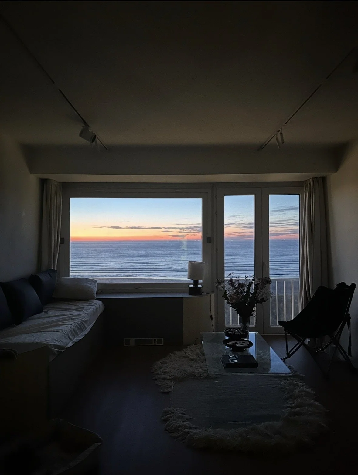 Living room with large window view of ocean at sunset, featuring a sofa, a side table with a lamp, a coffee table with flowers, and a lounge chair.