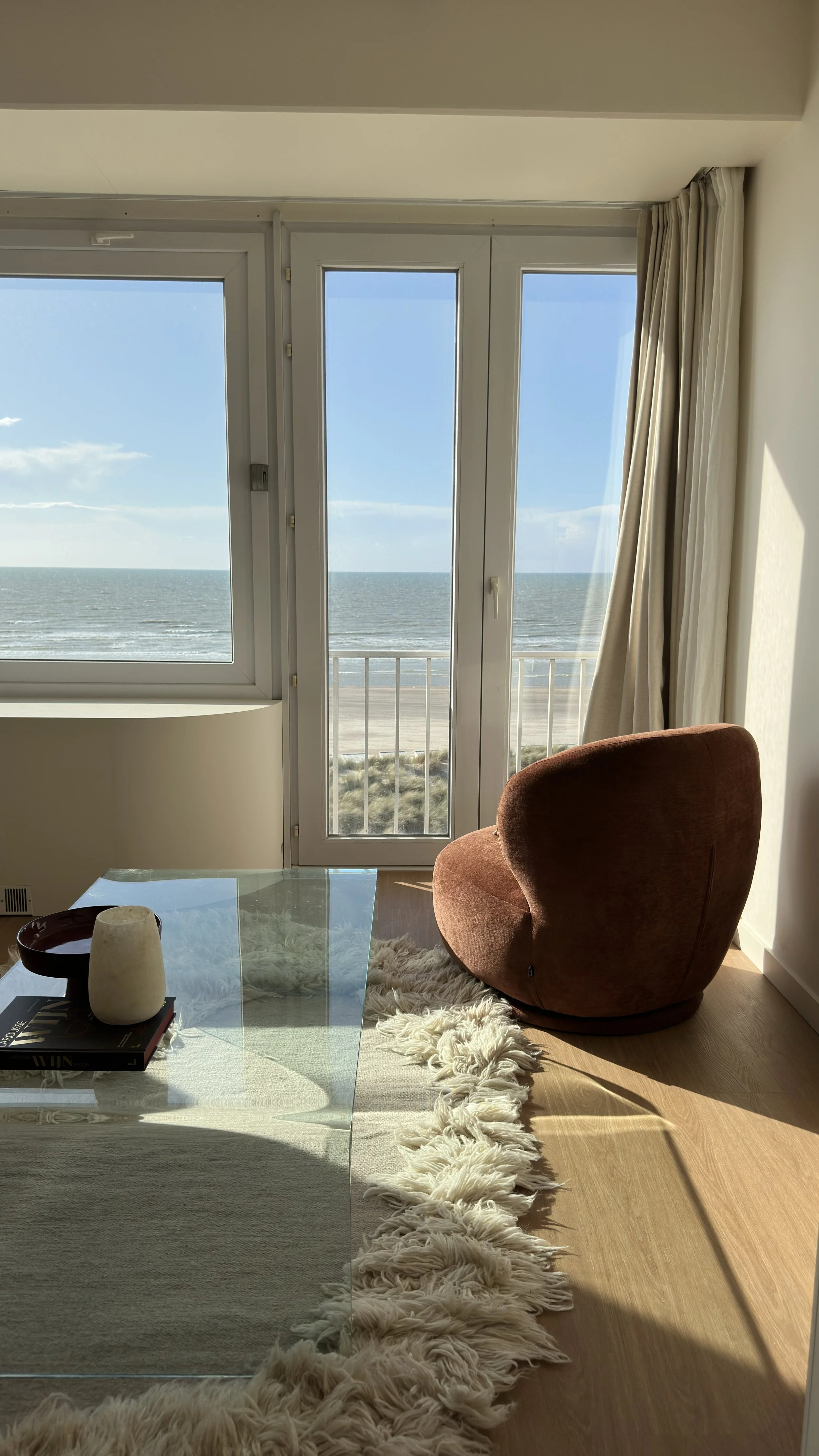 Room with ocean view through a glass door, brown armchair, beige curtains, glass coffee table with decorative items, cream rug.