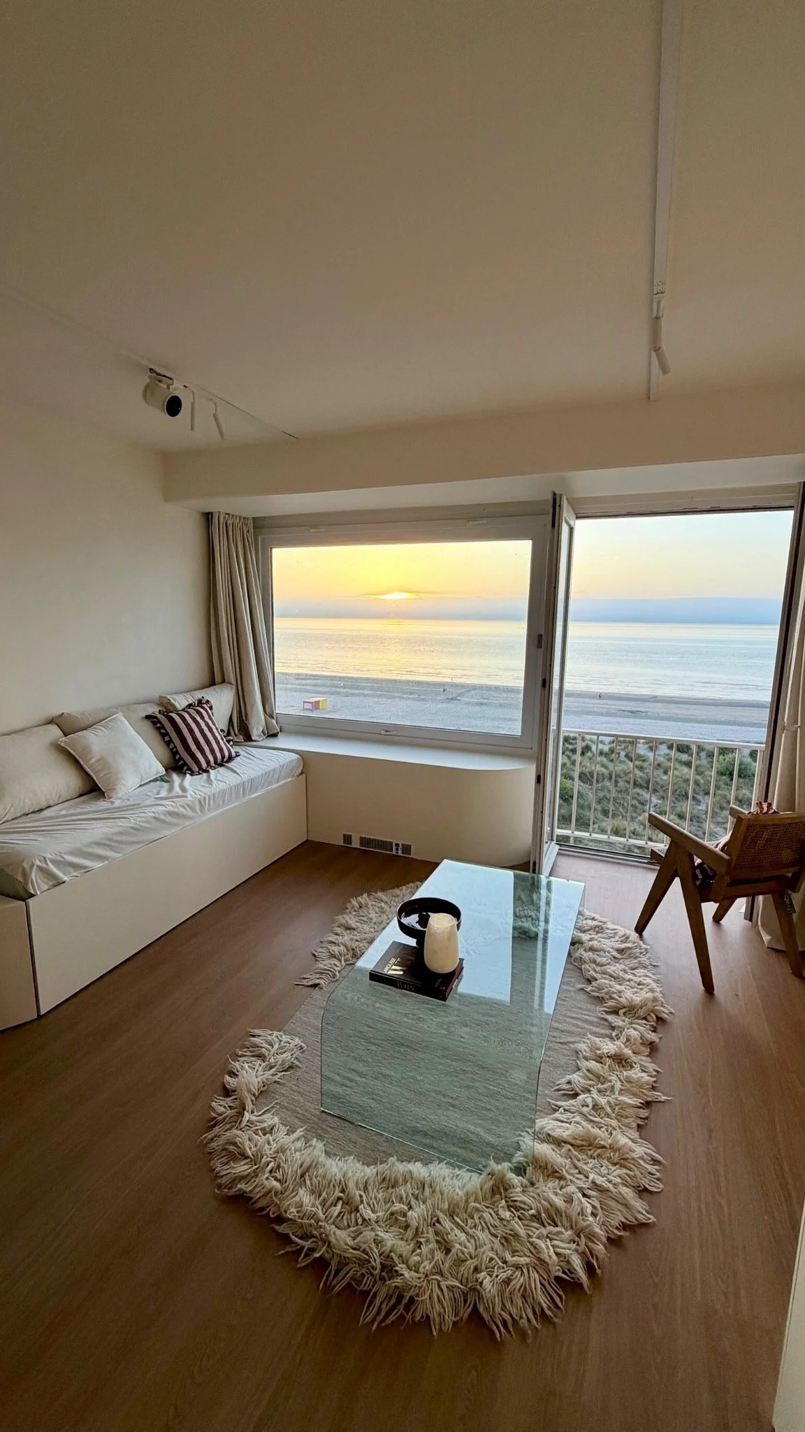 Living room with large window showing a beach sunset, with white sofa, glass coffee table, and a plush rug.