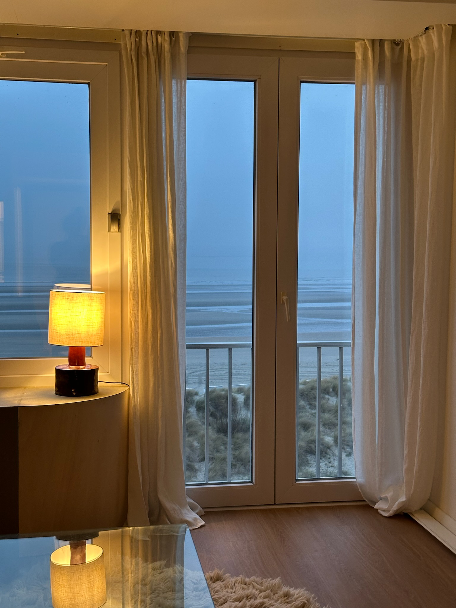A cozy room with a glass door leading to a beach view, white curtains, a table lamp with a beige shade, and a light-colored wooden floor.