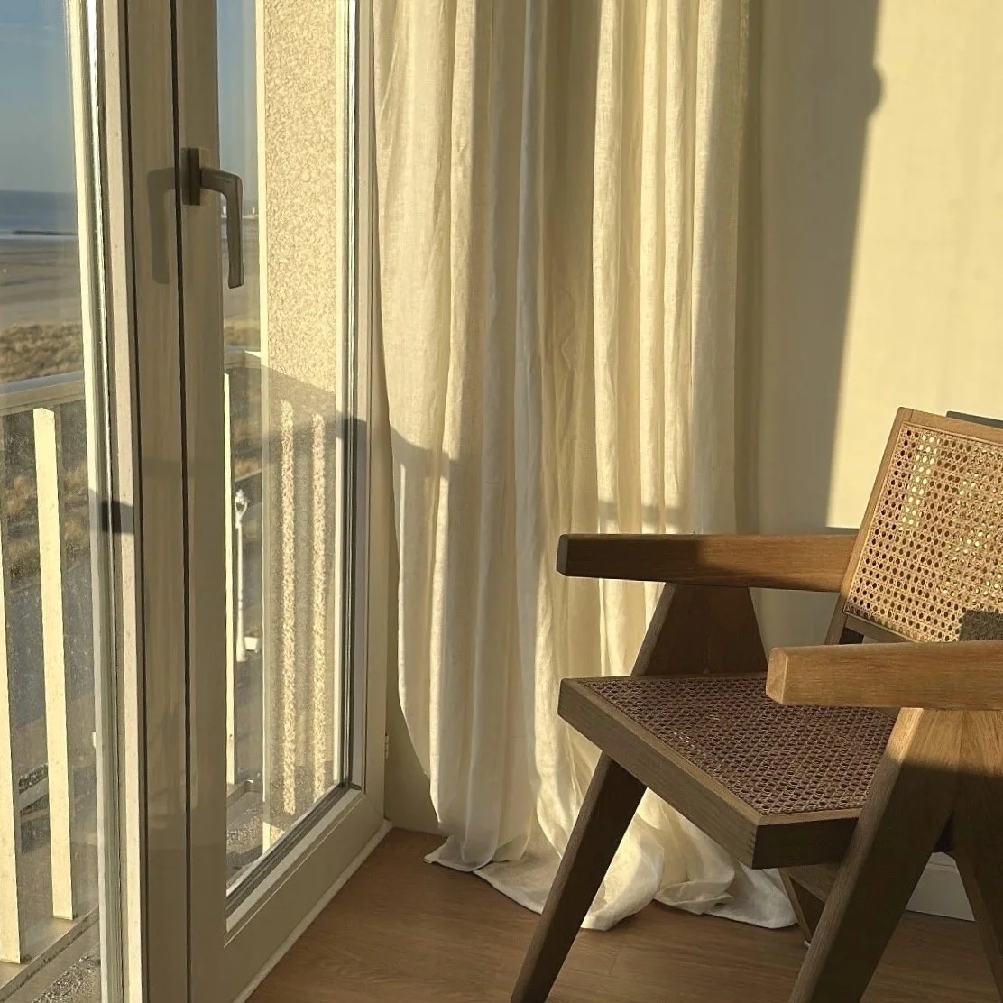 Sunlight coming through a glass sliding door with beige curtains, casting shadows on a wooden chair inside a room.