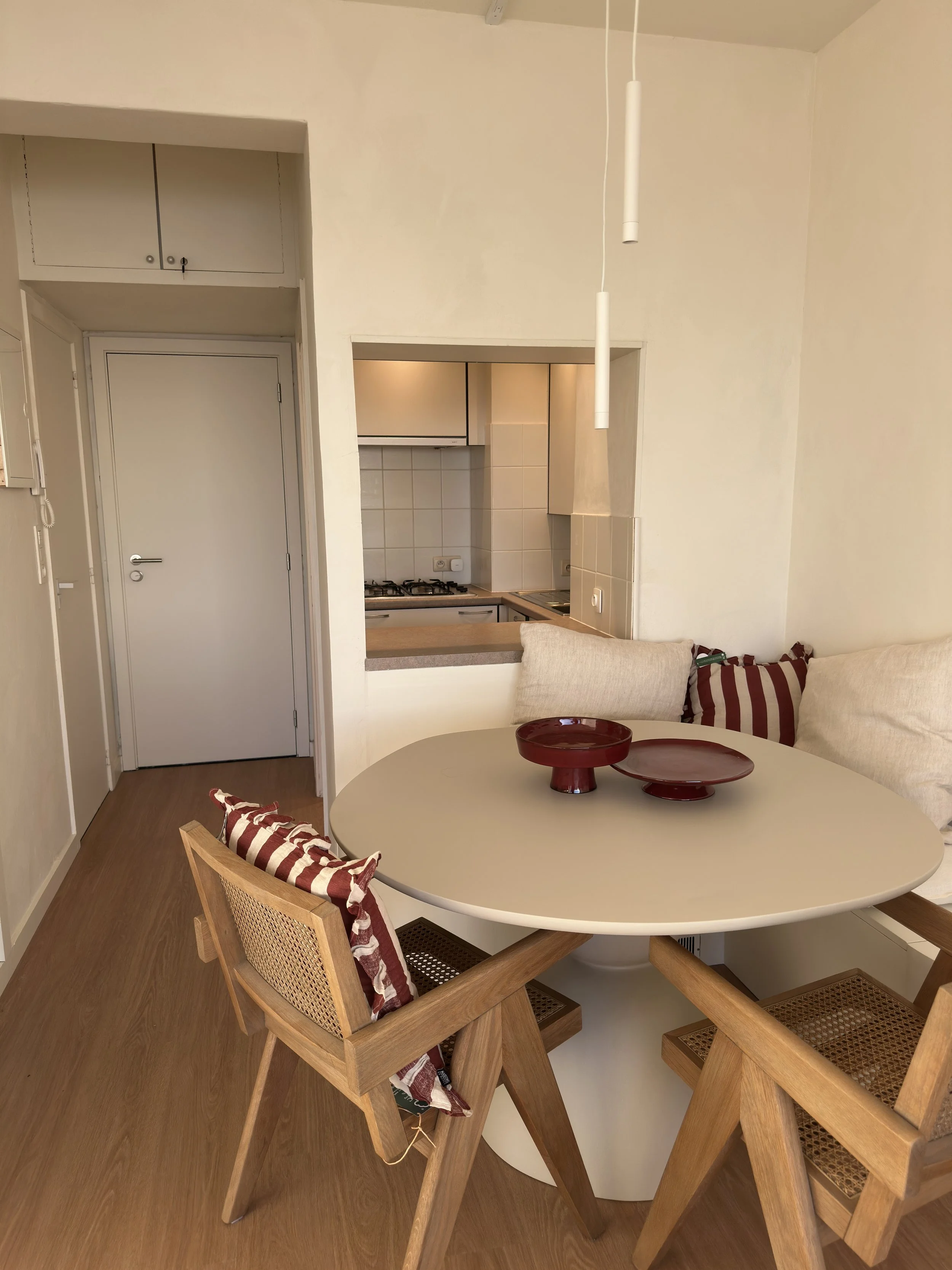A dining area with a wooden chair, a round table, and red bowls, next to a beige sofa with striped cushions, with a view into the kitchen through an open wall.
