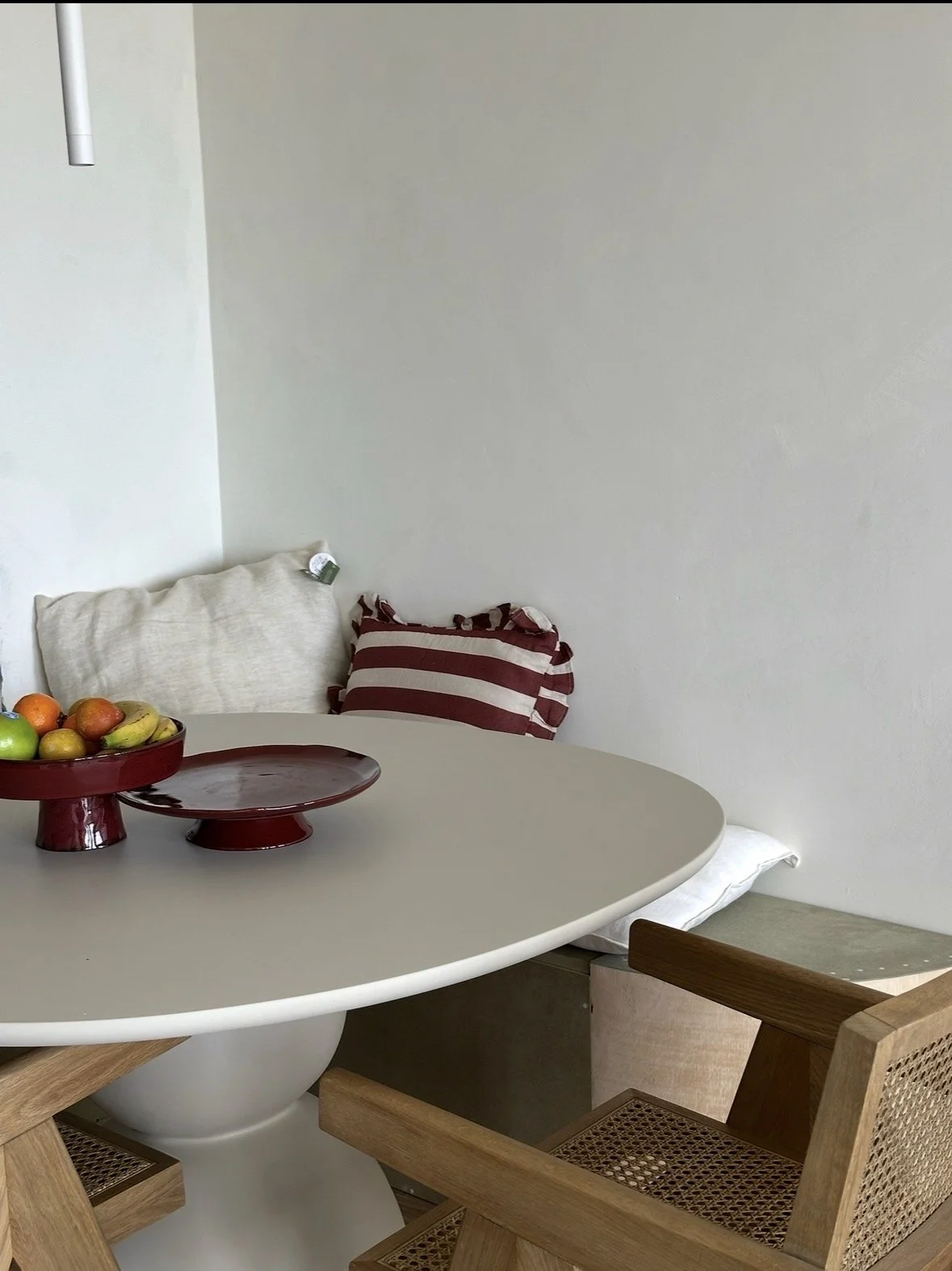 A modern dining area with a round white table, two wooden chairs with cane seats, and decorative bowls of fruit. The backdrop features a plain white wall with pillows resting against it.