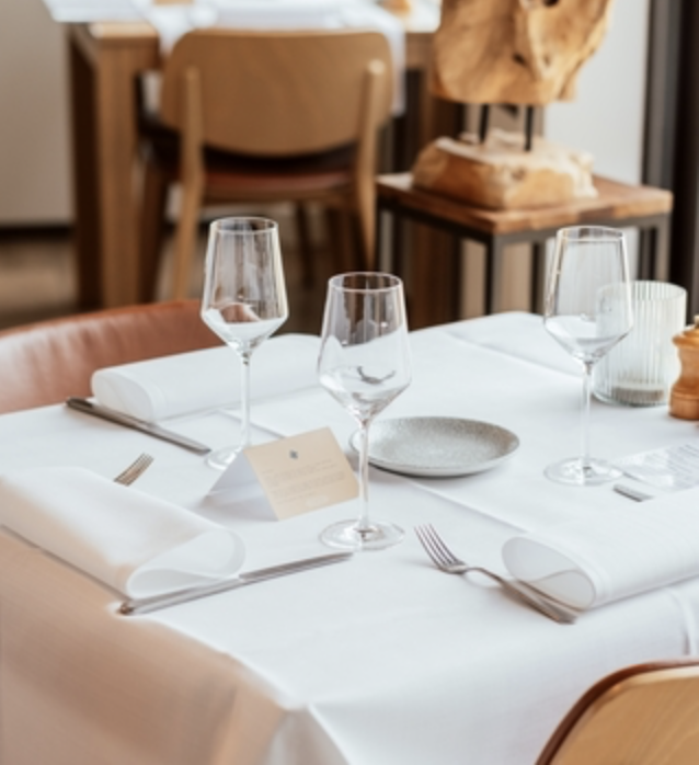 A elegantly set dining table with white tablecloth, three empty wine glasses, silverware, and a small menu card. In the background, there is a wooden chair and decorative items, including a sculpture on a wooden stand.