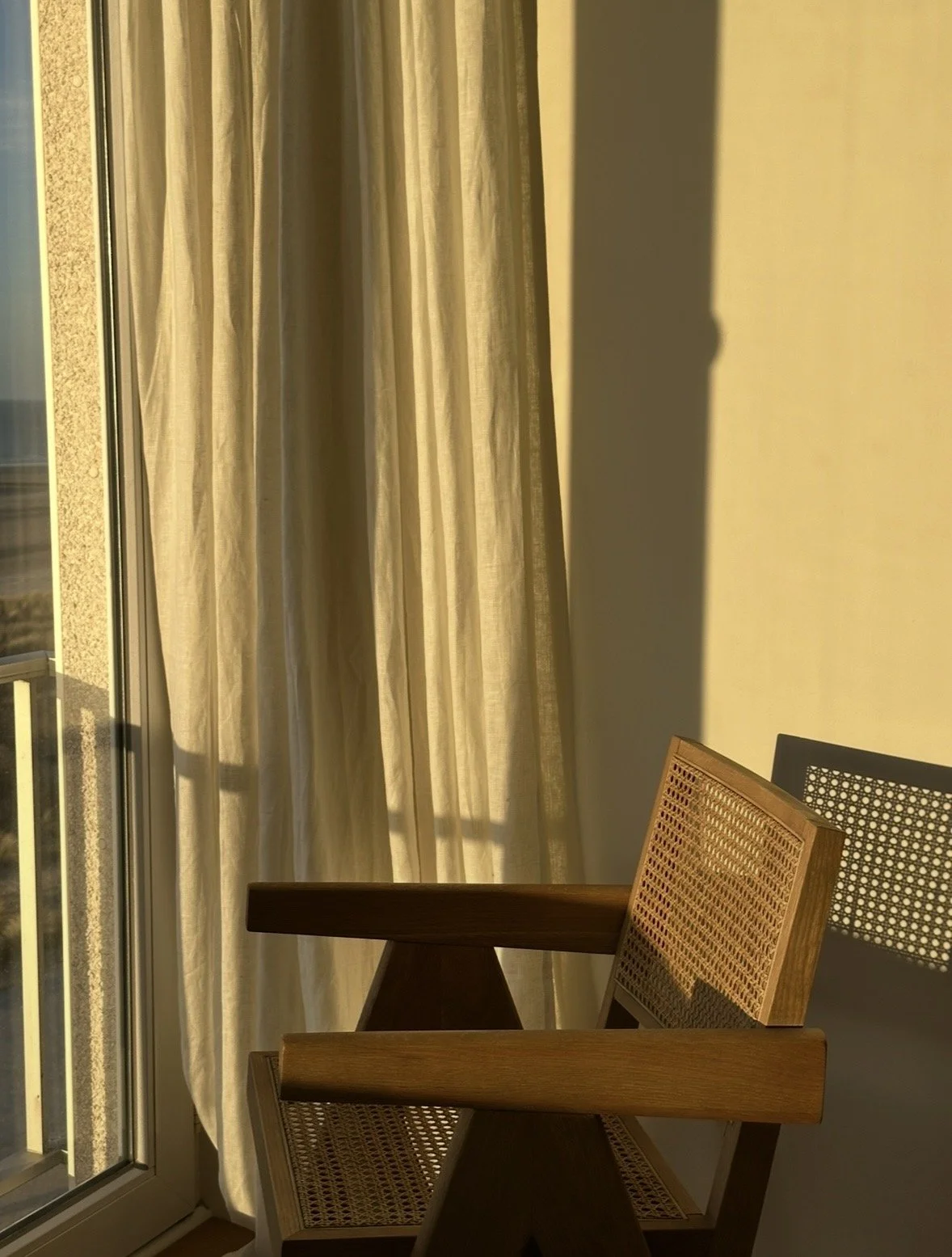 A wooden chair with cane backrest near a sliding glass door with beige curtains, sunlight casting shadows, and a yellow wall.