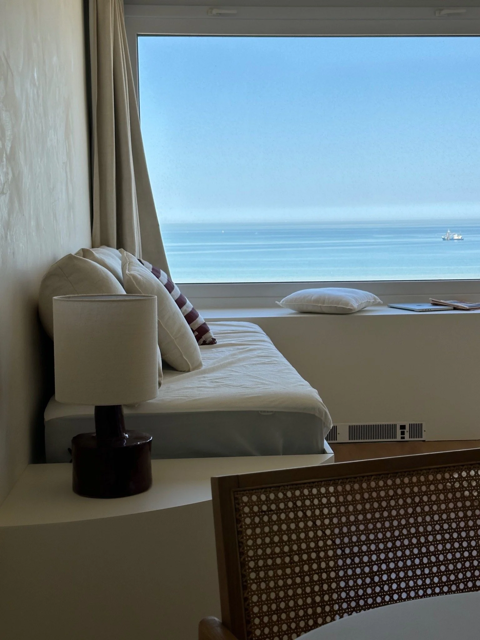 Room with a large window showing the ocean, a ship in the distance, beige curtains, a white bed with pillows, a wooden chair with a decorative back, and a small table with a lamp and books.