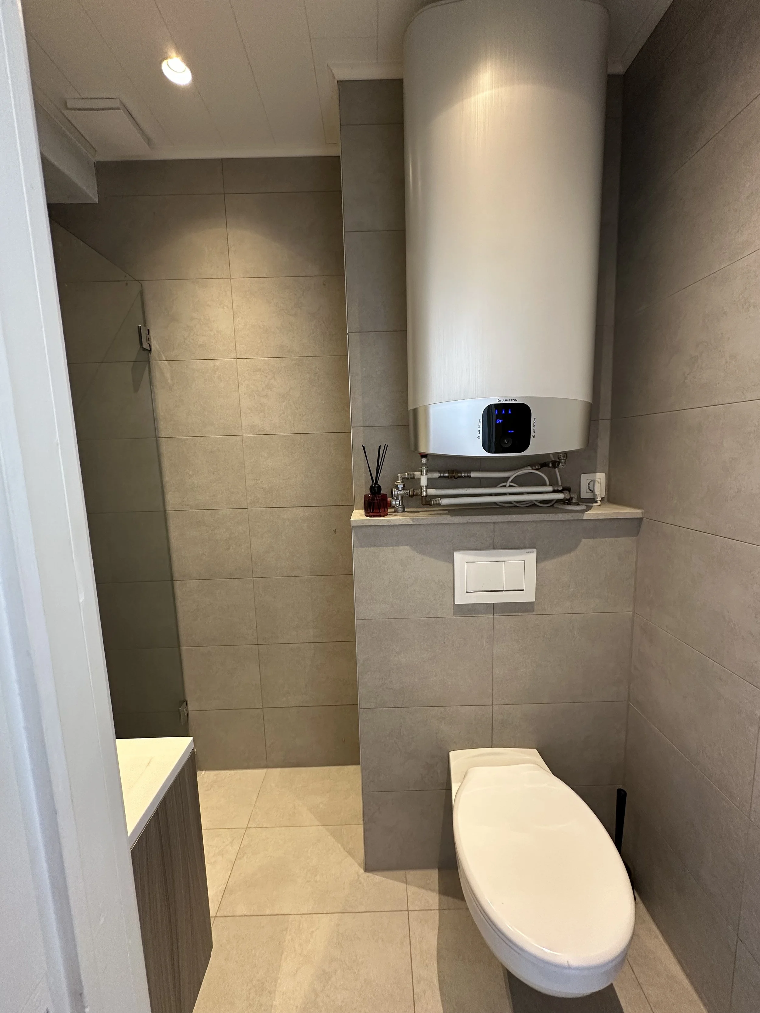 A modern bathroom with beige tiled walls and floor, a wall-mounted toilet, an electric water heater, a small sink with a vanity, and a reed diffuser on the countertop.