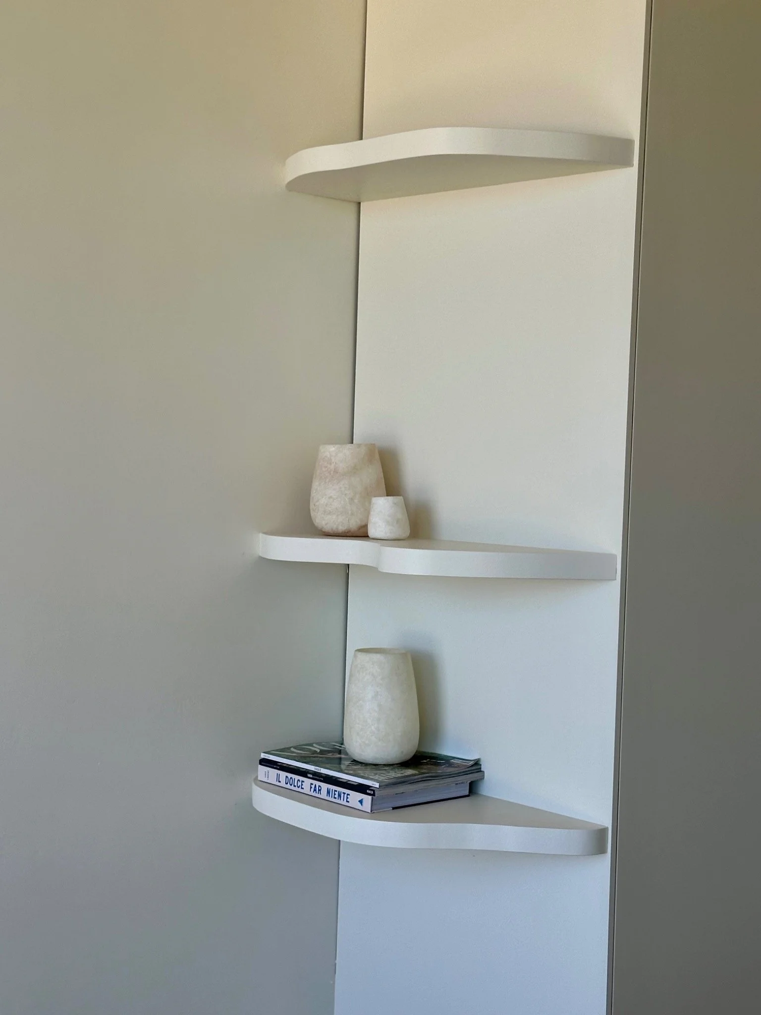 White wall-mounted corner shelf with three vases on it, stacked vertically, with a few magazines or books underneath.