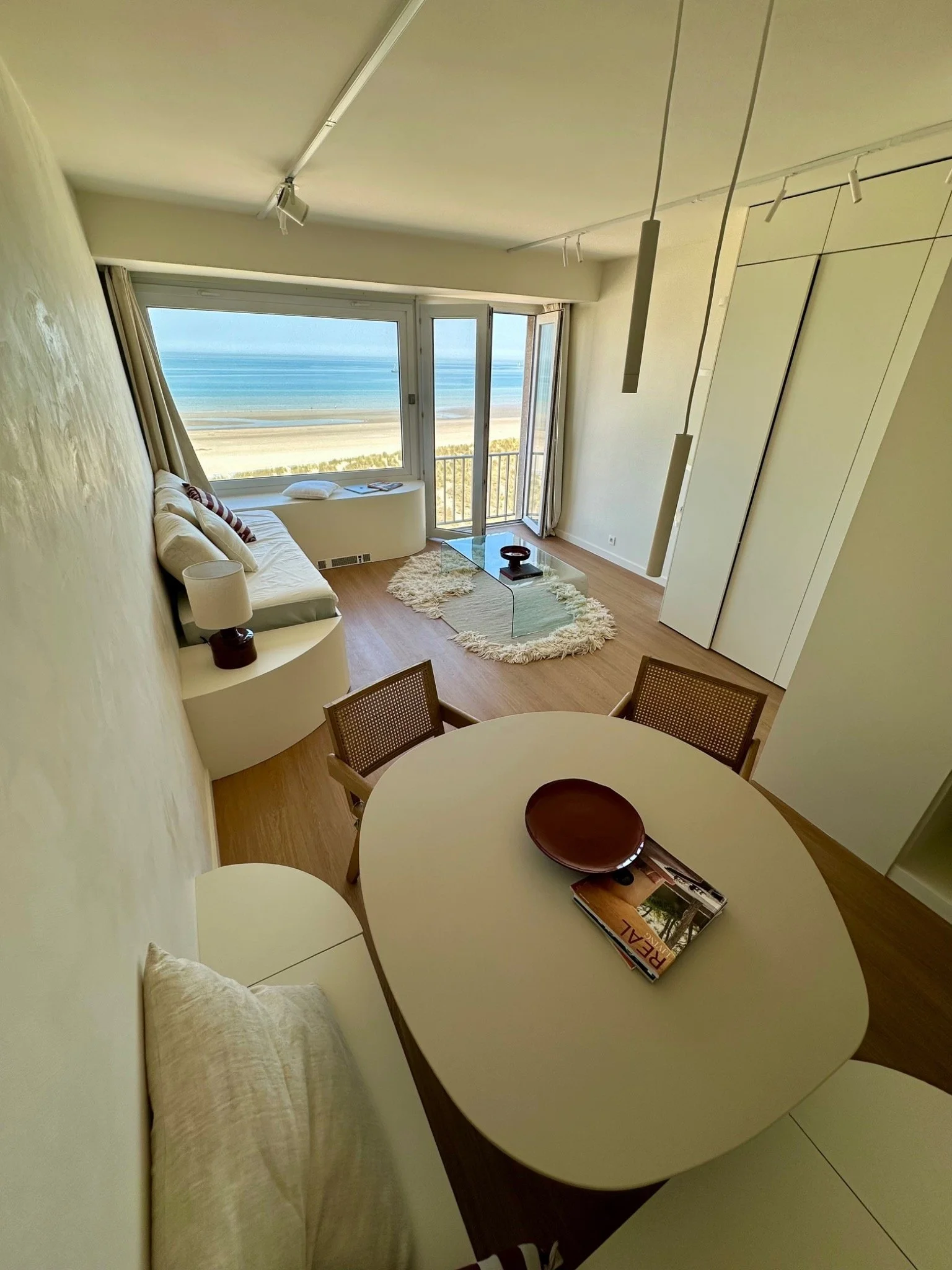 A living room with a large window and balcony overlooking a beach and ocean, with a white sofa, a glass coffee table, and a small round dining table with chairs.