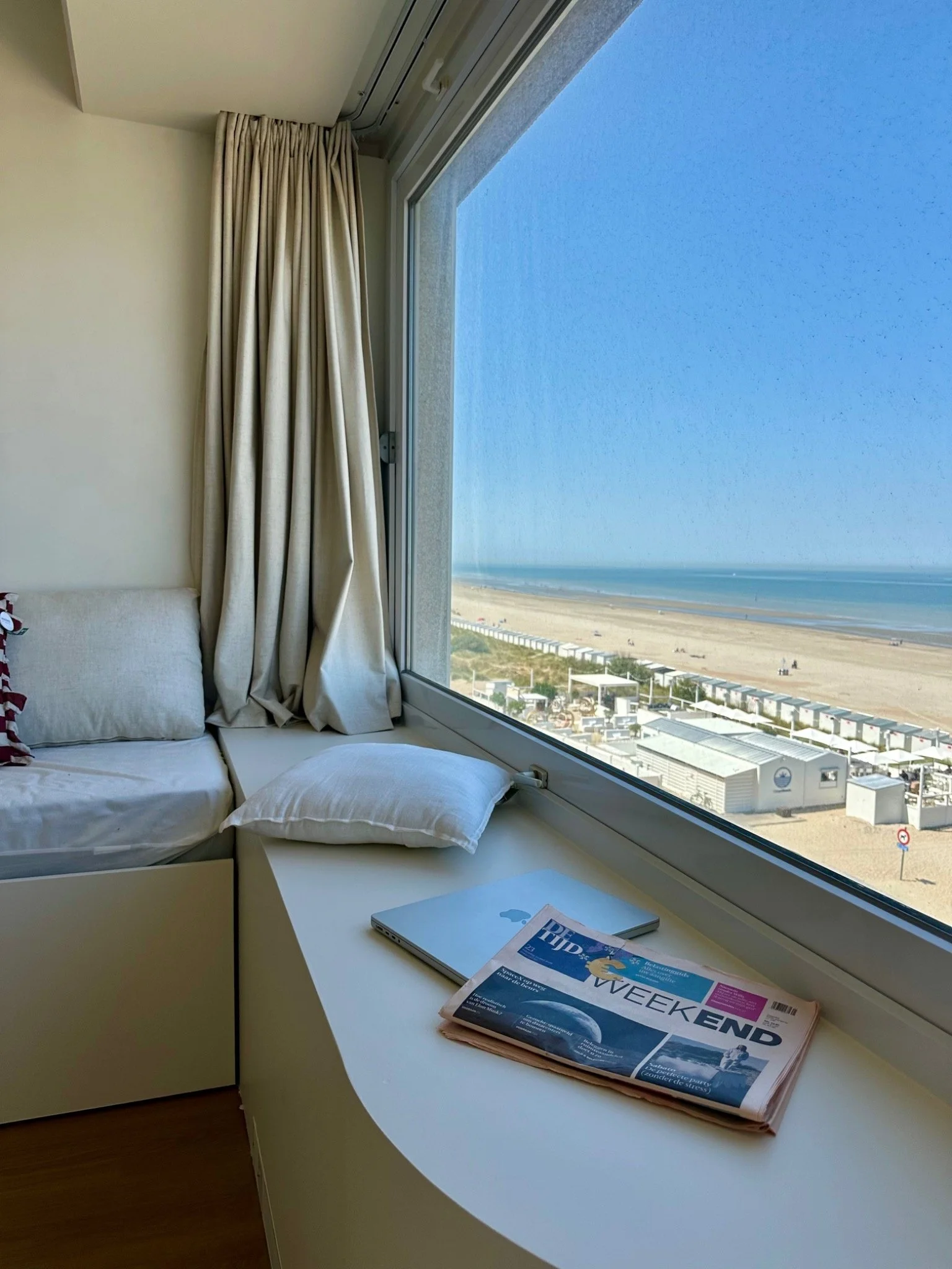 A white desk with a smartphone and a newspaper titled 'Weekend' placed on it, situated next to a large window with a view of a beach and buildings below. Cream-colored curtains partially cover the window, and there are pillows on a sofa or bed near t