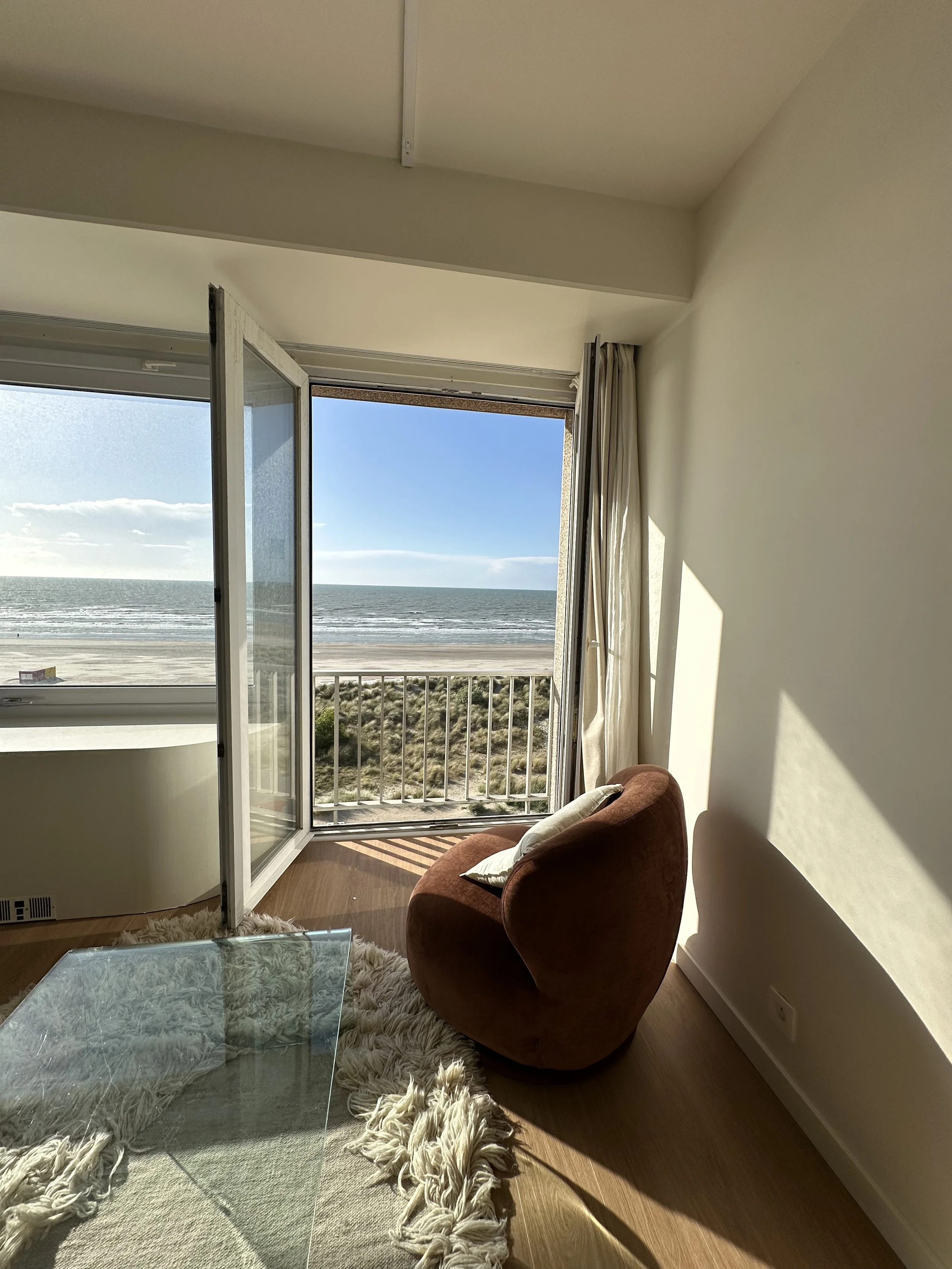 A cozy living room with a large open window showing a beach and ocean view, a brown armchair with a white pillow, a glass coffee table, a fluffy white rug, and sunlight casting shadows on the cream-colored walls.