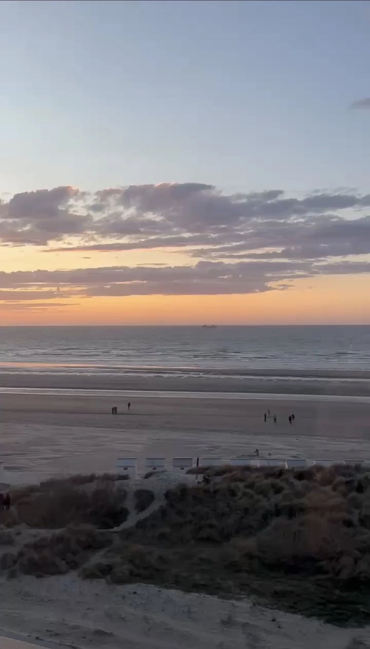 Beach scene during sunset with a colorful sky, calm ocean, and a few small groups of people walking along the sand.