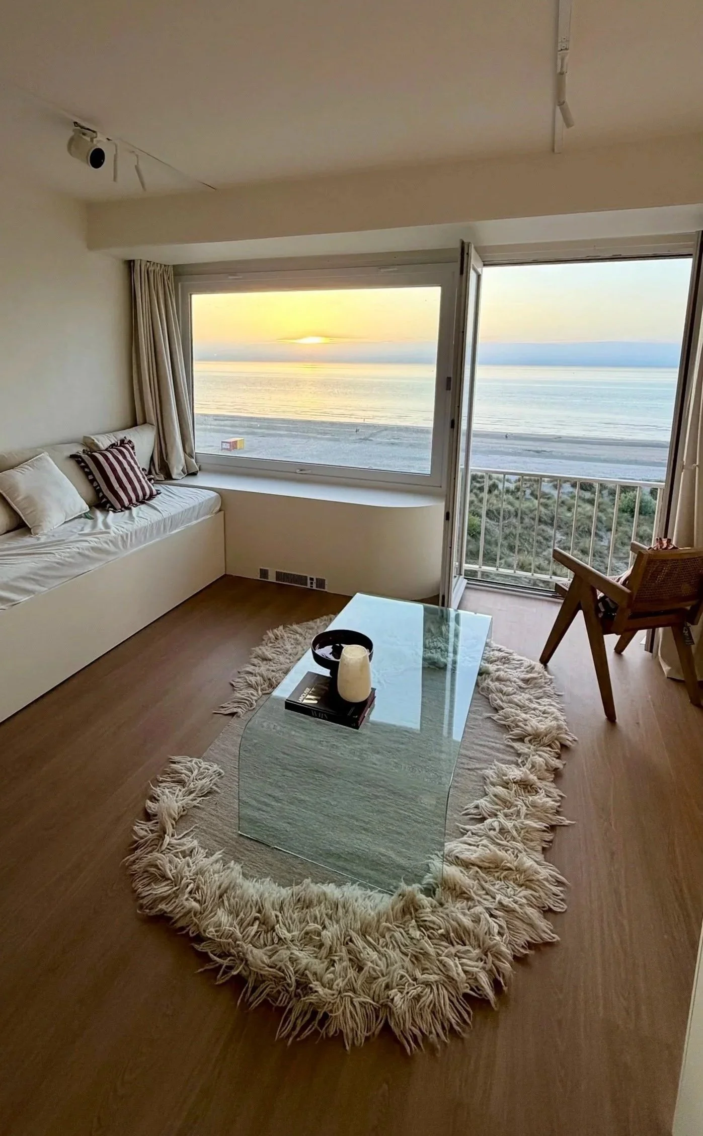 Living room with a large window overlooking the ocean during sunset, white sofa with striped pillows, glass coffee table with books and candles, shaggy rug, wooden chair, and curtains.