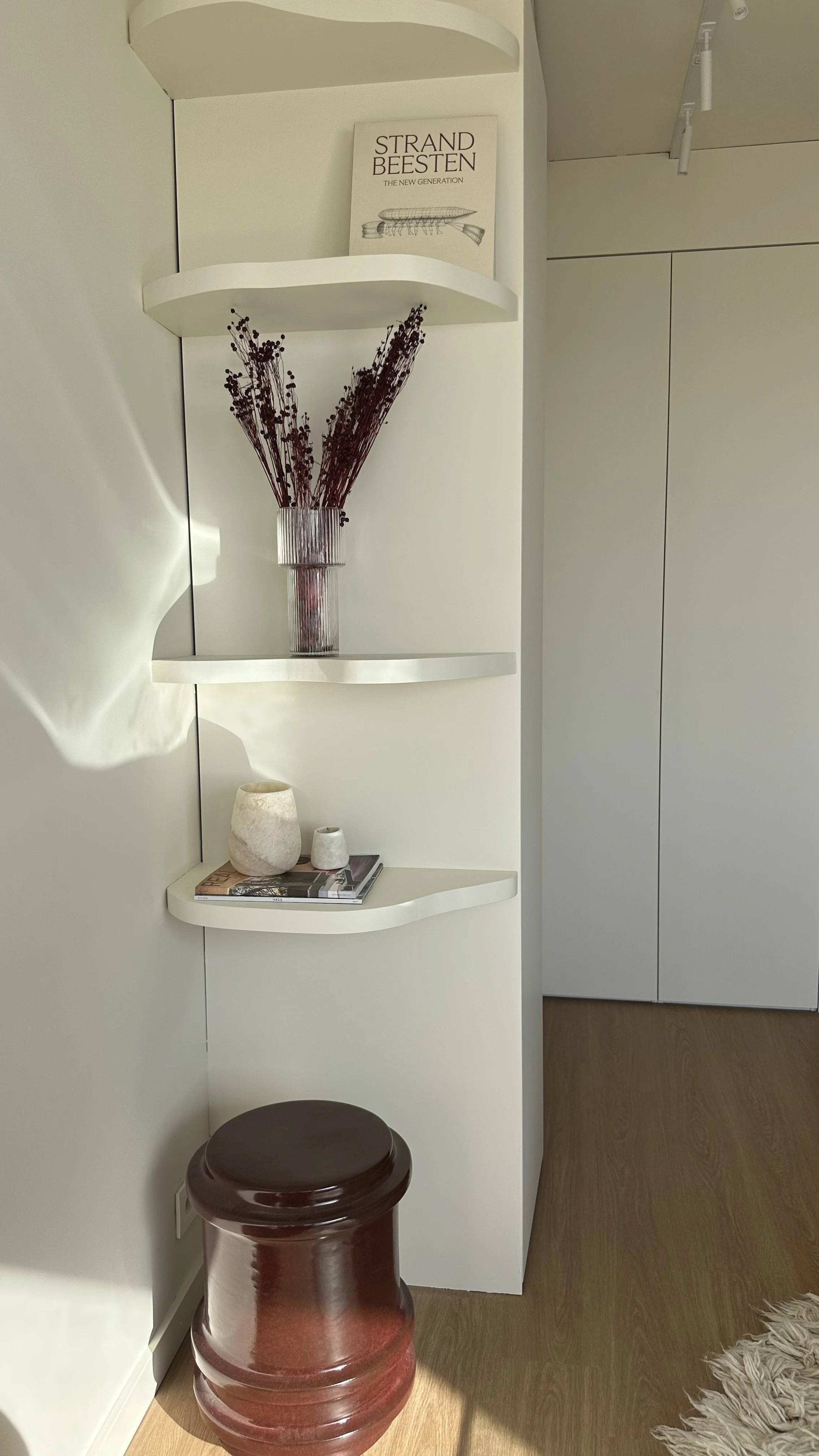 Interior of a modern room with a curved white wall shelf holding decorative items including a book, a tall vase with dried flowers, and two smaller objects on a magazine. There is a dark red ceramic stool in front of the shelf and a shaggy cream-colo